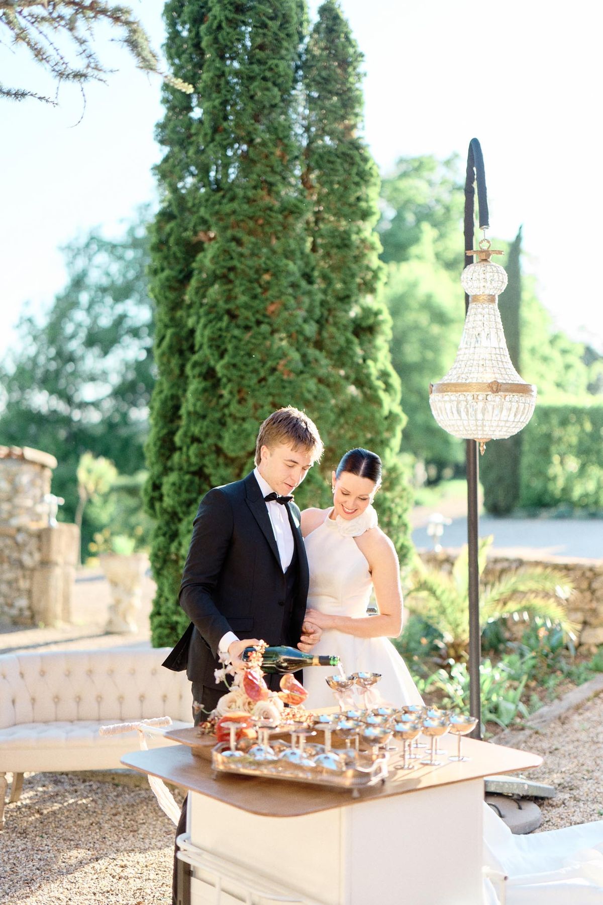 Couple pouring champagne at outdoor bar with crystal chandelier and tufted sofa on chateau terrace