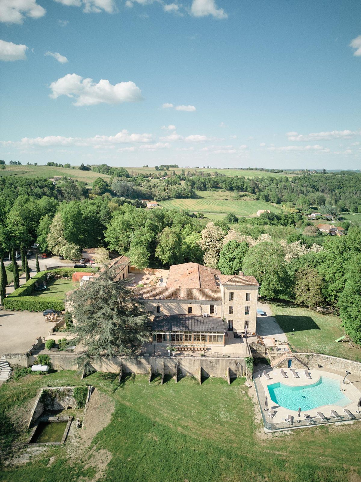 Aerial French estate with terracotta roofs, glass orangery, kidney pool, and cedar tree in countryside