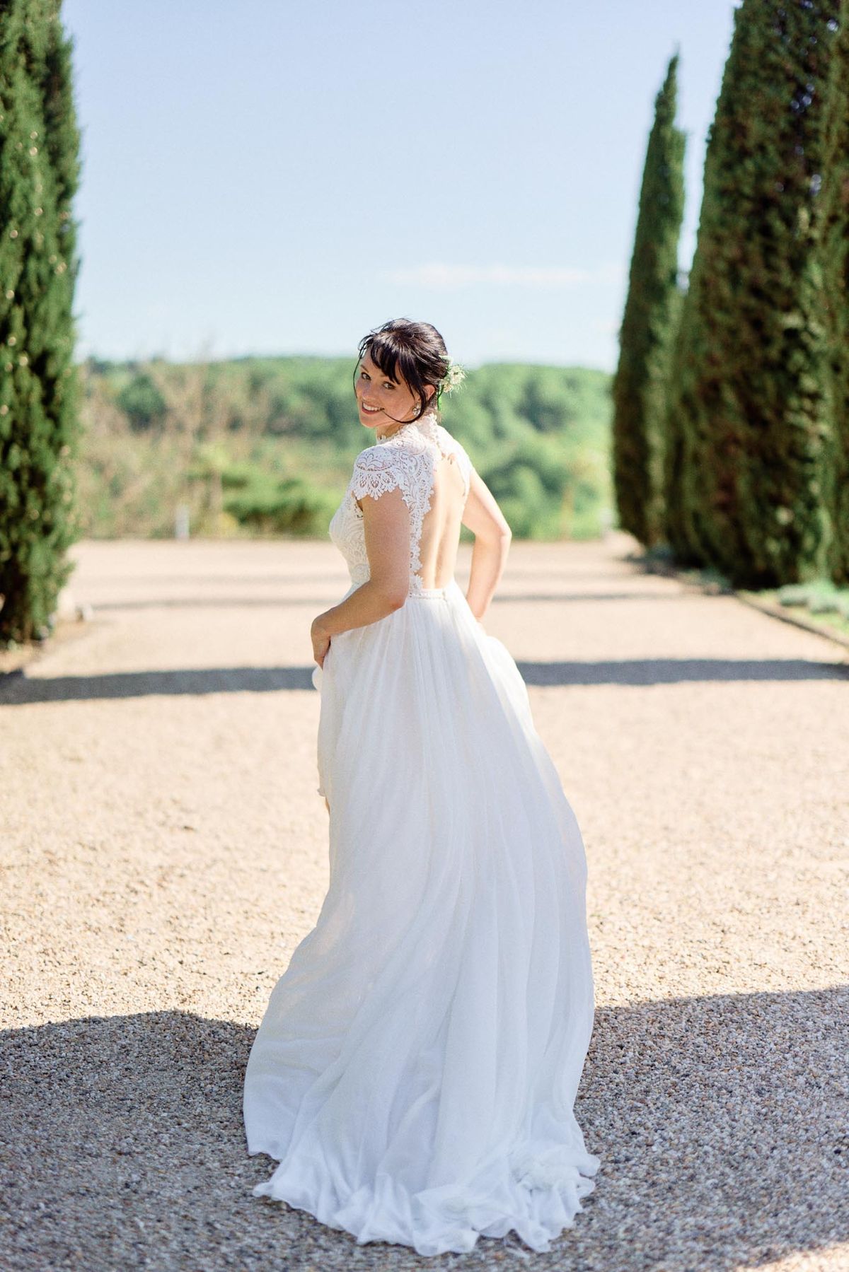 Bride looking over shoulder on cypress-lined gravel allee wearing lace cap-sleeve open-back gown