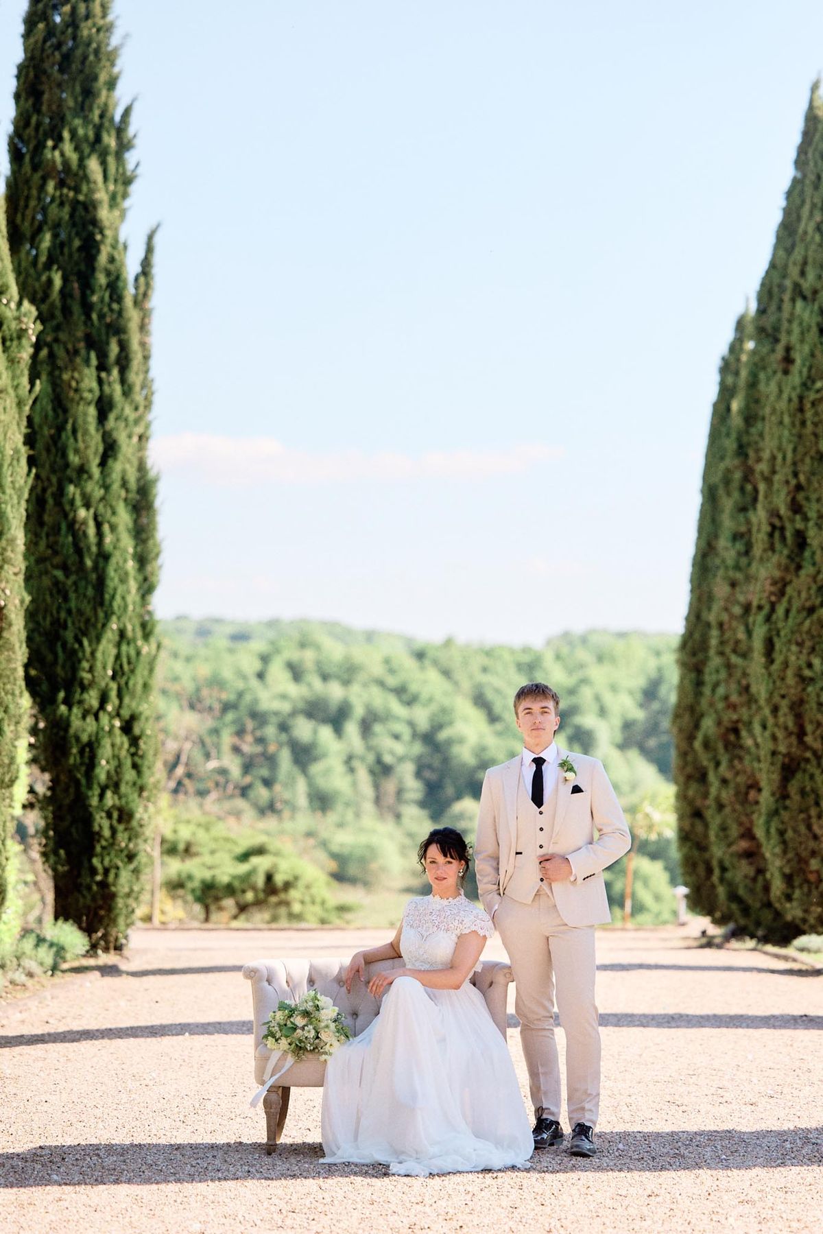 Bride seated in lace ball gown and groom in sand suit on settee along cypress-lined allee at French chateau