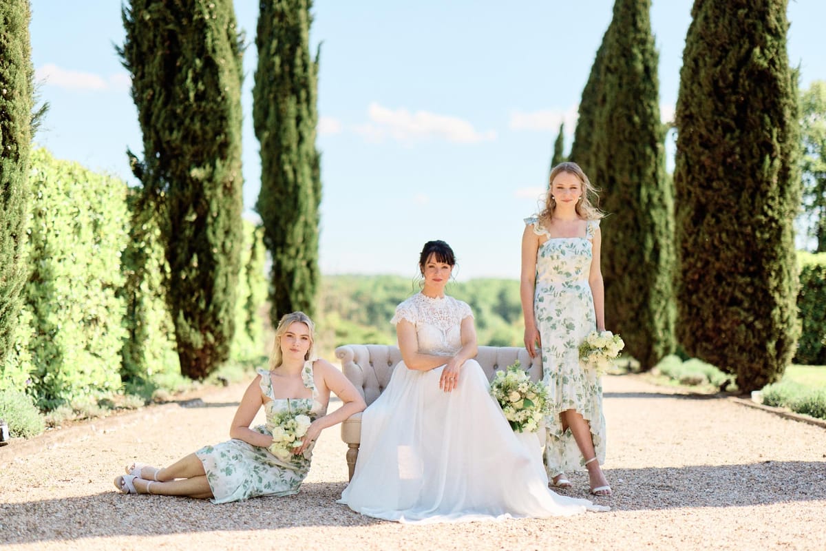 Bride on tufted settee with two bridesmaids in sage botanical print dresses on cypress-lined gravel allee