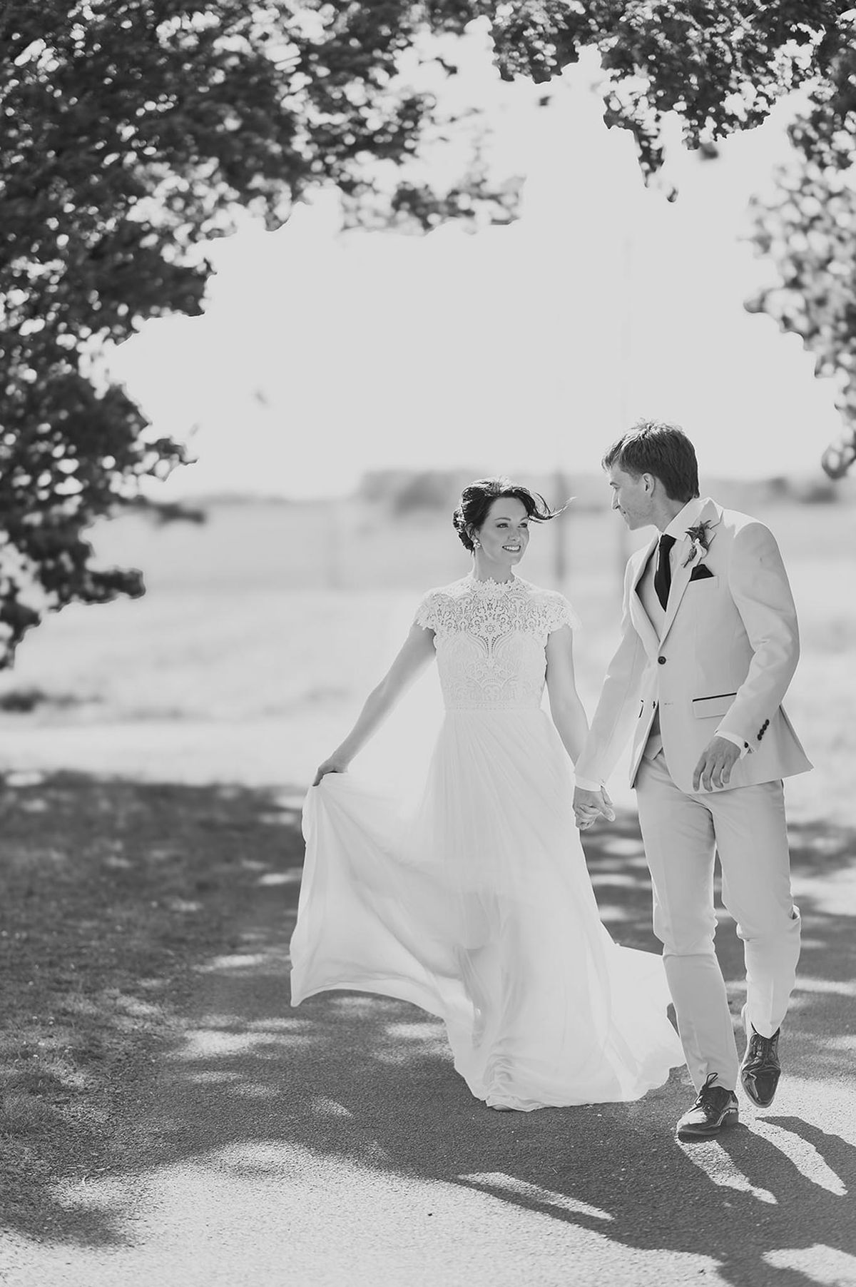 Black and white portrait of bride and groom walking hand in hand on tree-lined path