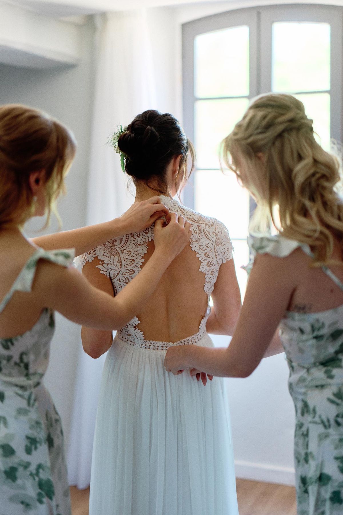Two bridesmaids fastening the lace back of the bride's gown in a bright room with arched French windows