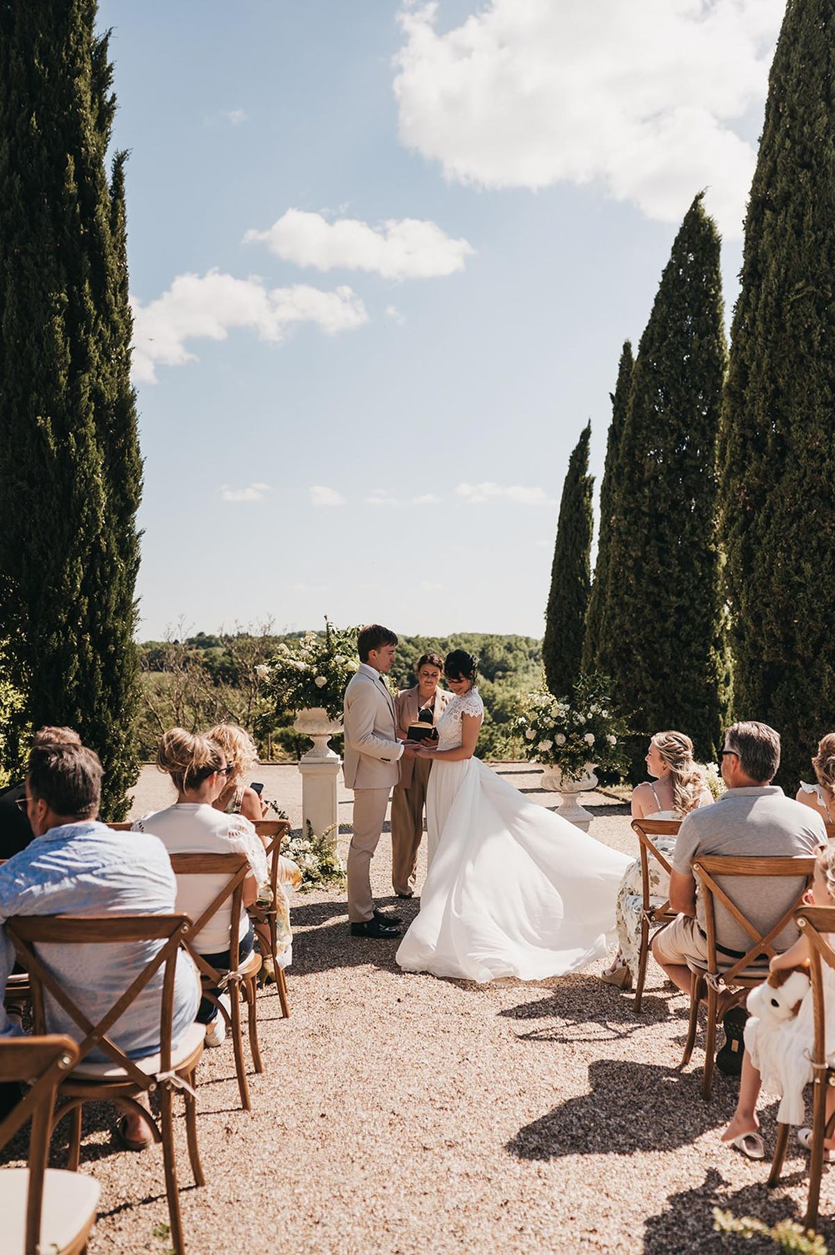 Intimate outdoor ceremony between cypress trees with white floral urns and cross-back chairs on gravel
