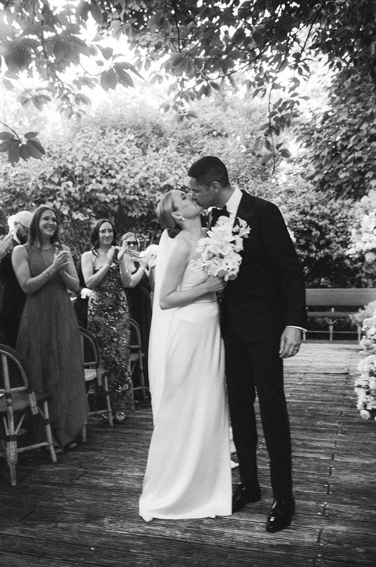 Black and white kiss in the aisle as guests applaud beneath the trees at Restaurant l'Ile