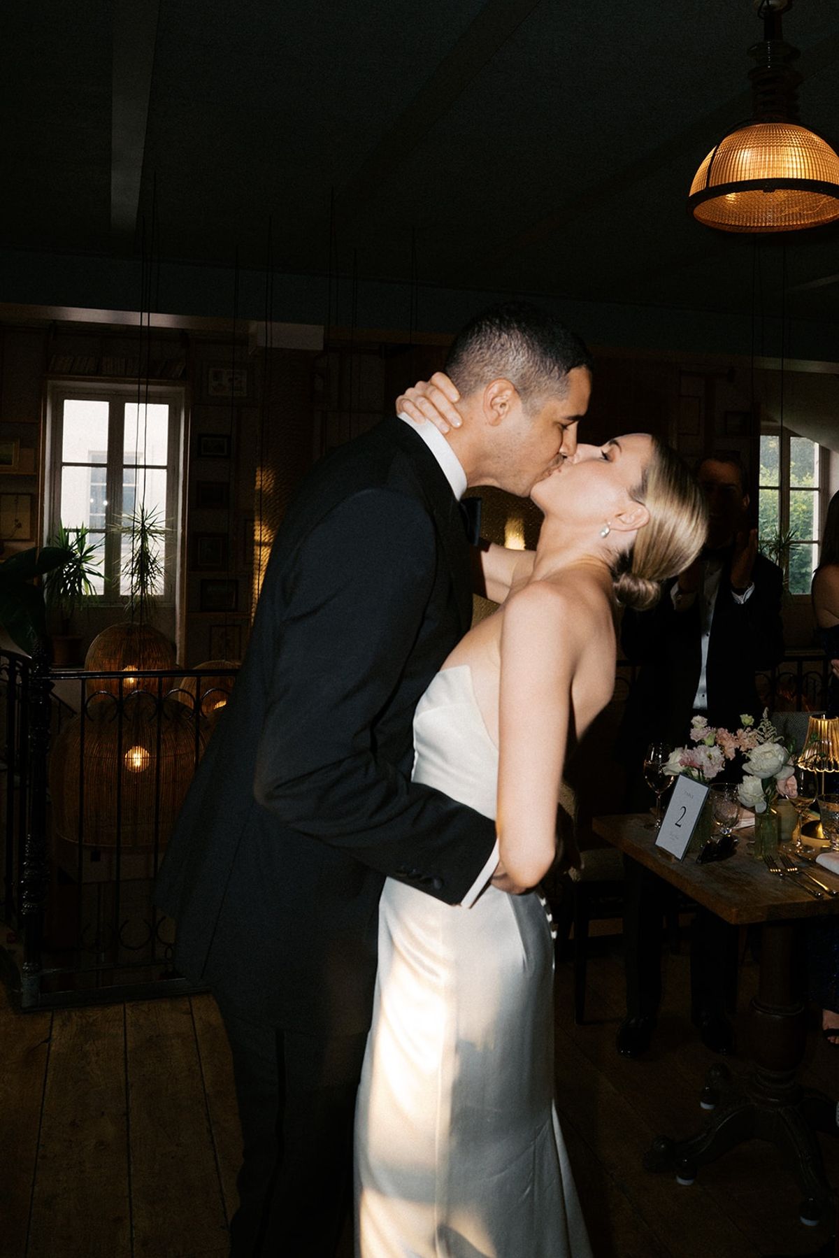 Couple kissing at the reception table beneath a brass pendant lamp