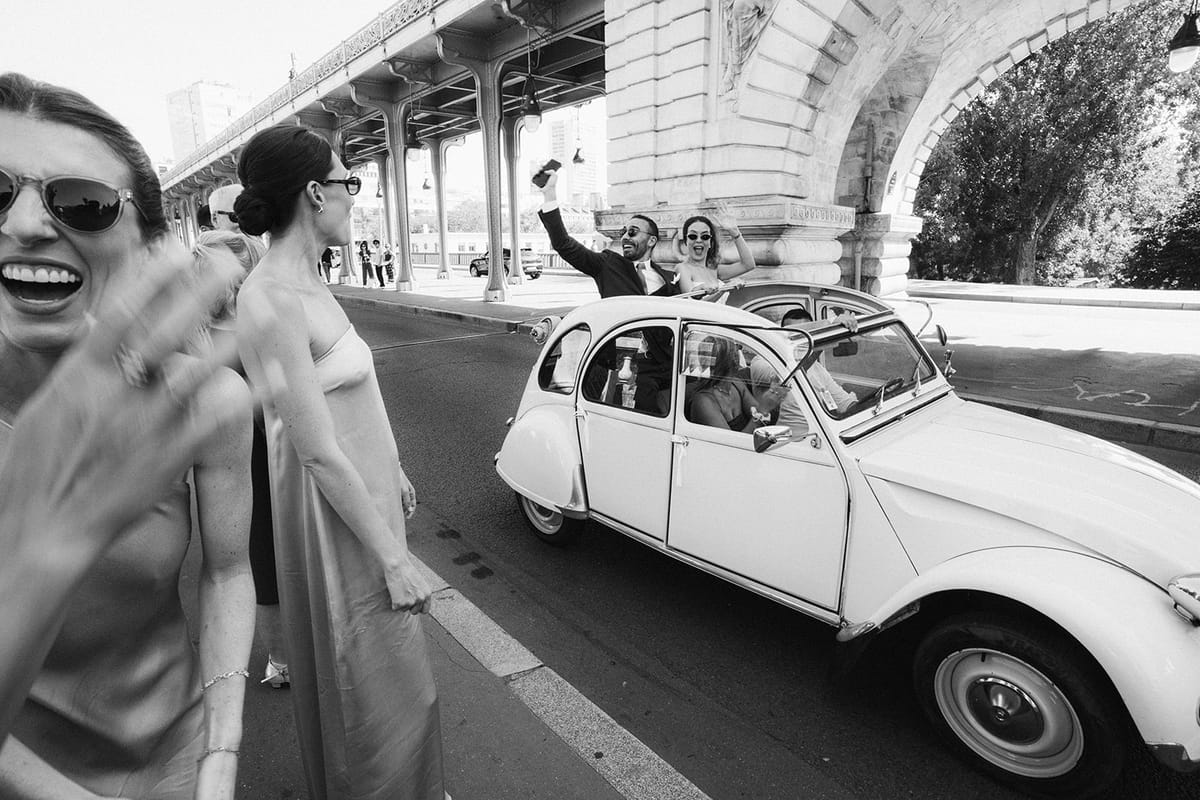 Black and white shot of guests waving at a vintage Citroen 2CV beneath a Paris bridge