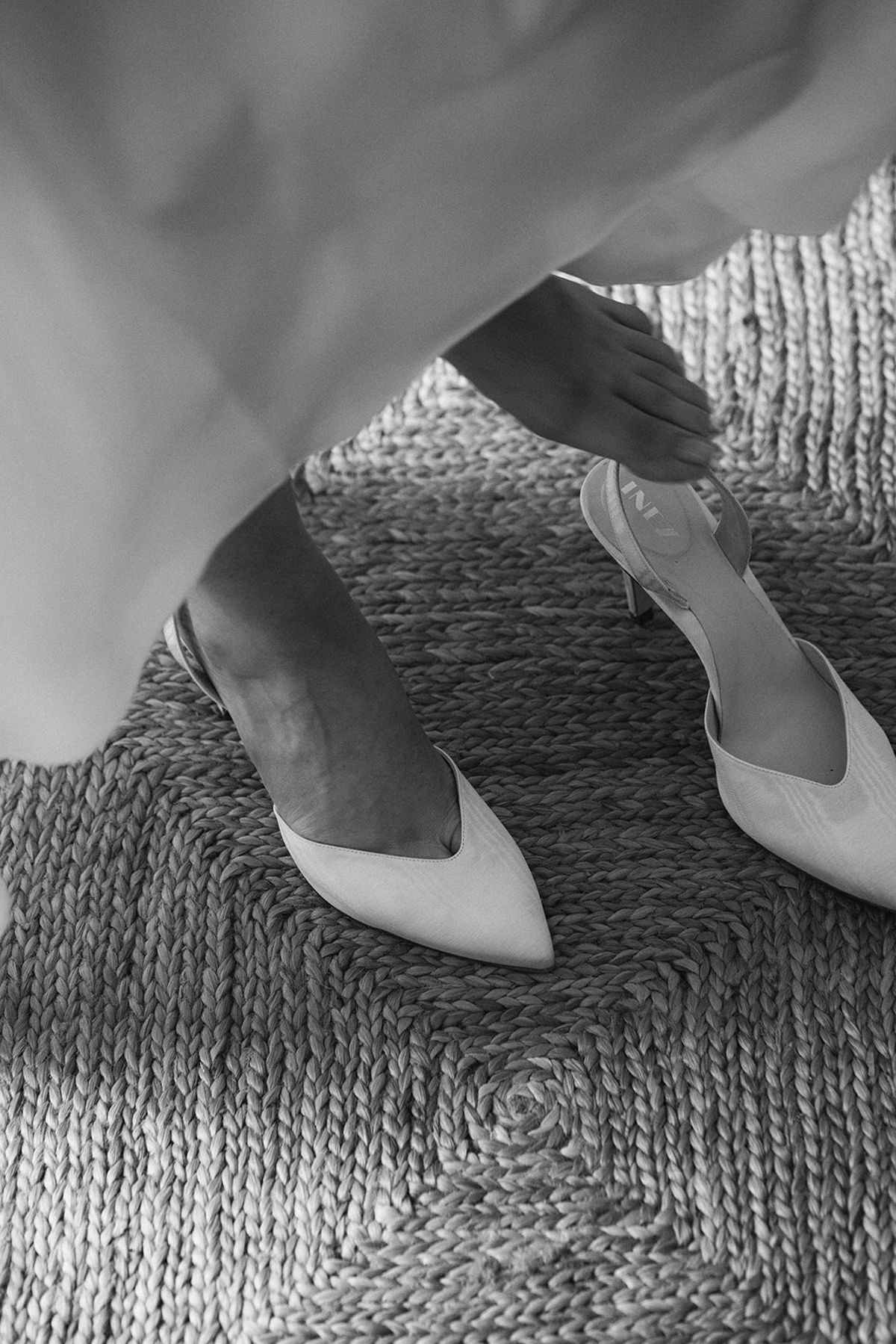 Black and white shot of the bride stepping into pointed slingbacks on a woven jute rug