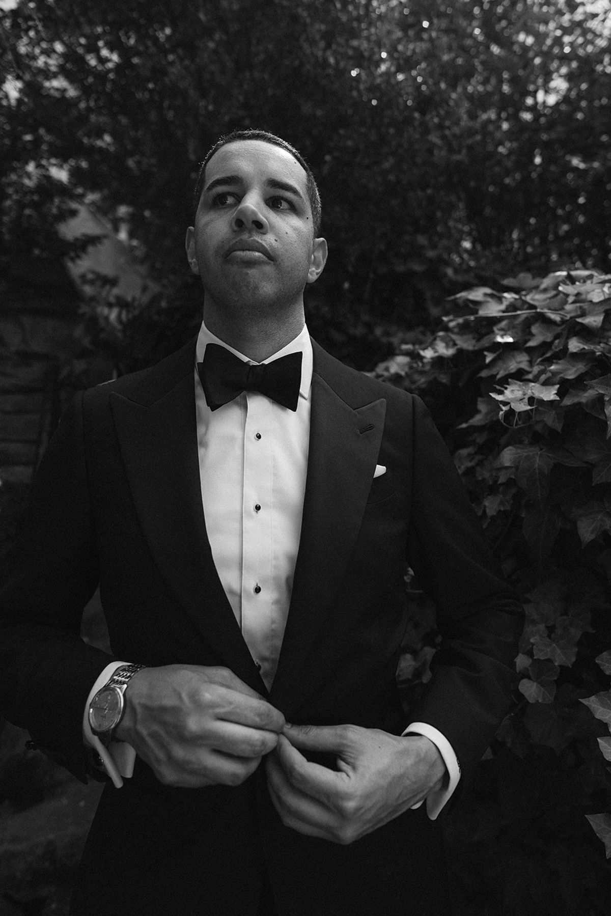 Black and white portrait of the groom buttoning his tuxedo against an ivy-covered wall