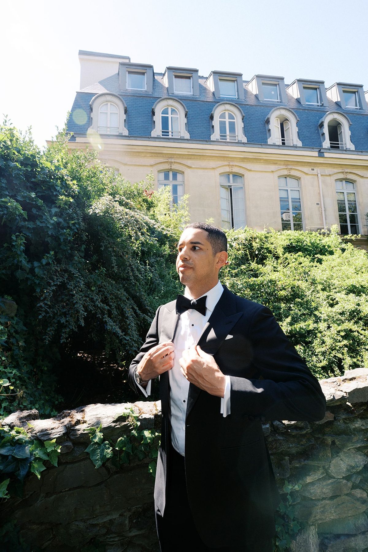 Groom buttoning his tuxedo on a stone garden wall with a Mansard-roofed Paris building behind