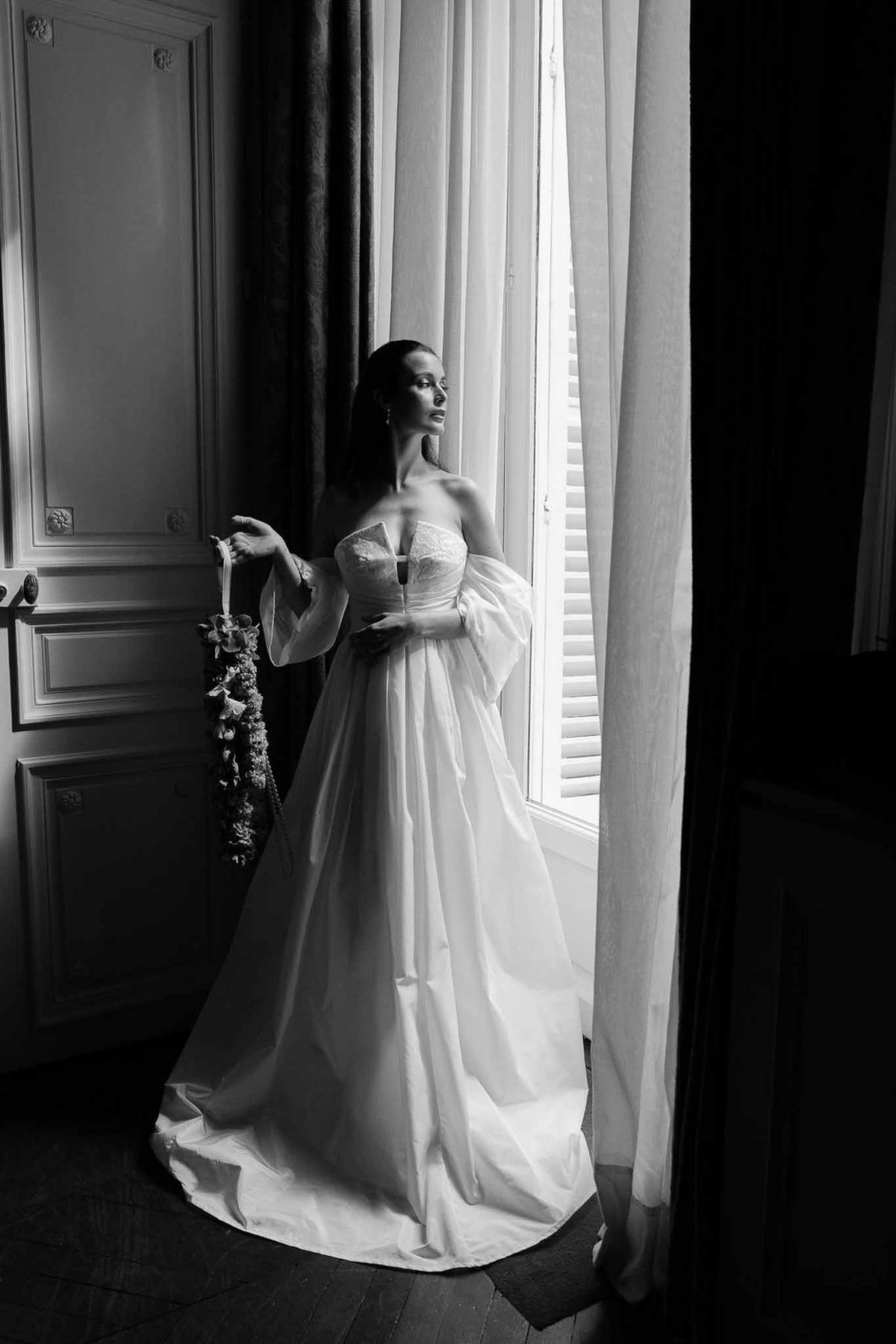 Bride in lace gown with billowing sleeves holds hanging floral beside tall window in paneled salon in B&W
