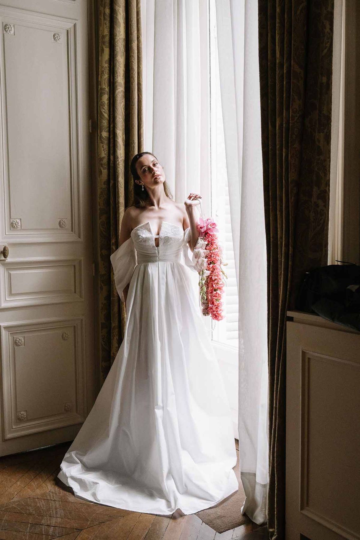 Bride in strapless gown with sheer sleeves holding cascading coral sweet pea bouquet by chateau window