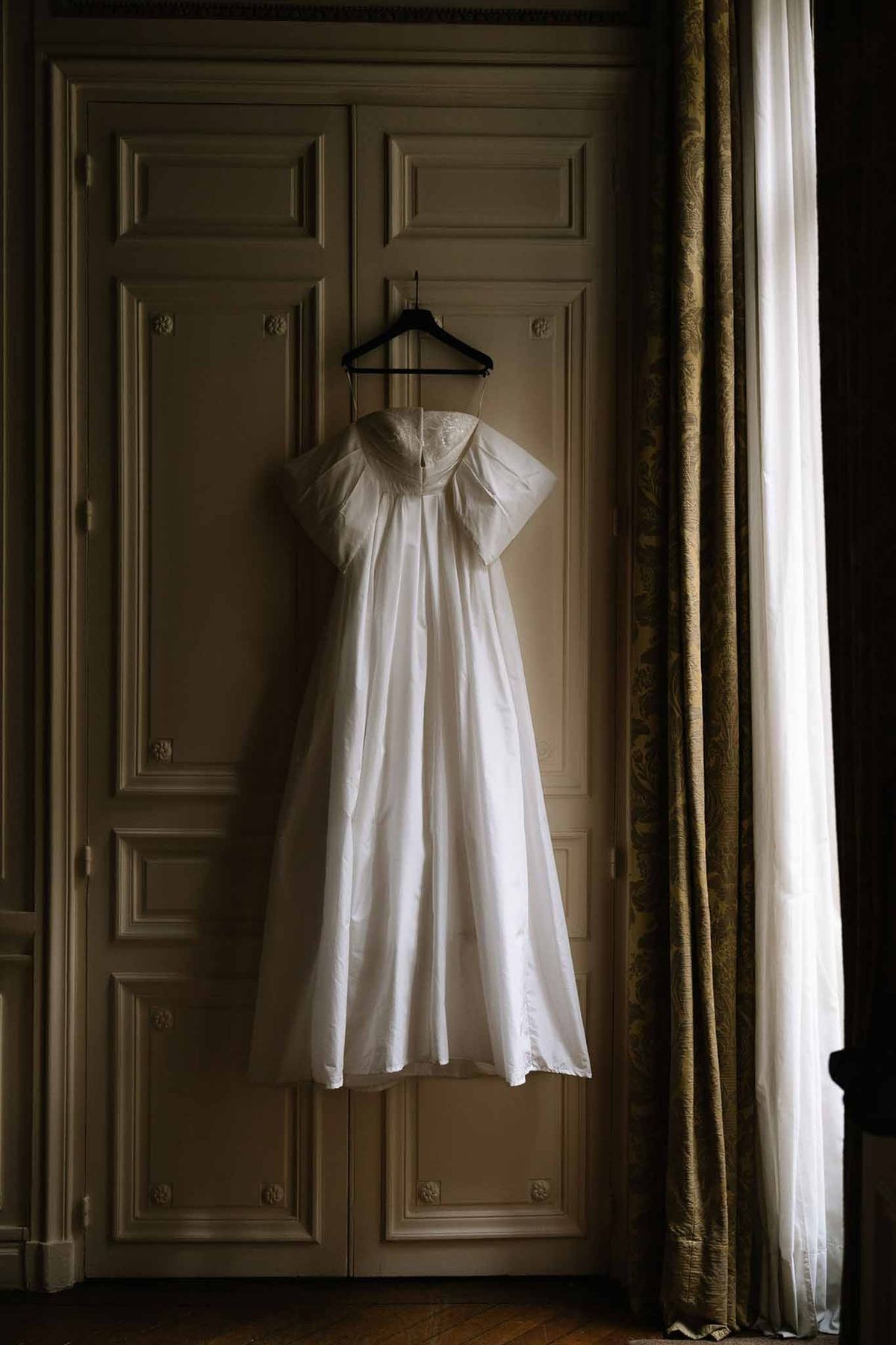 Ivory strapless wedding gown with sheer lace overskirt hanging on ornate paneled doors in a French interior