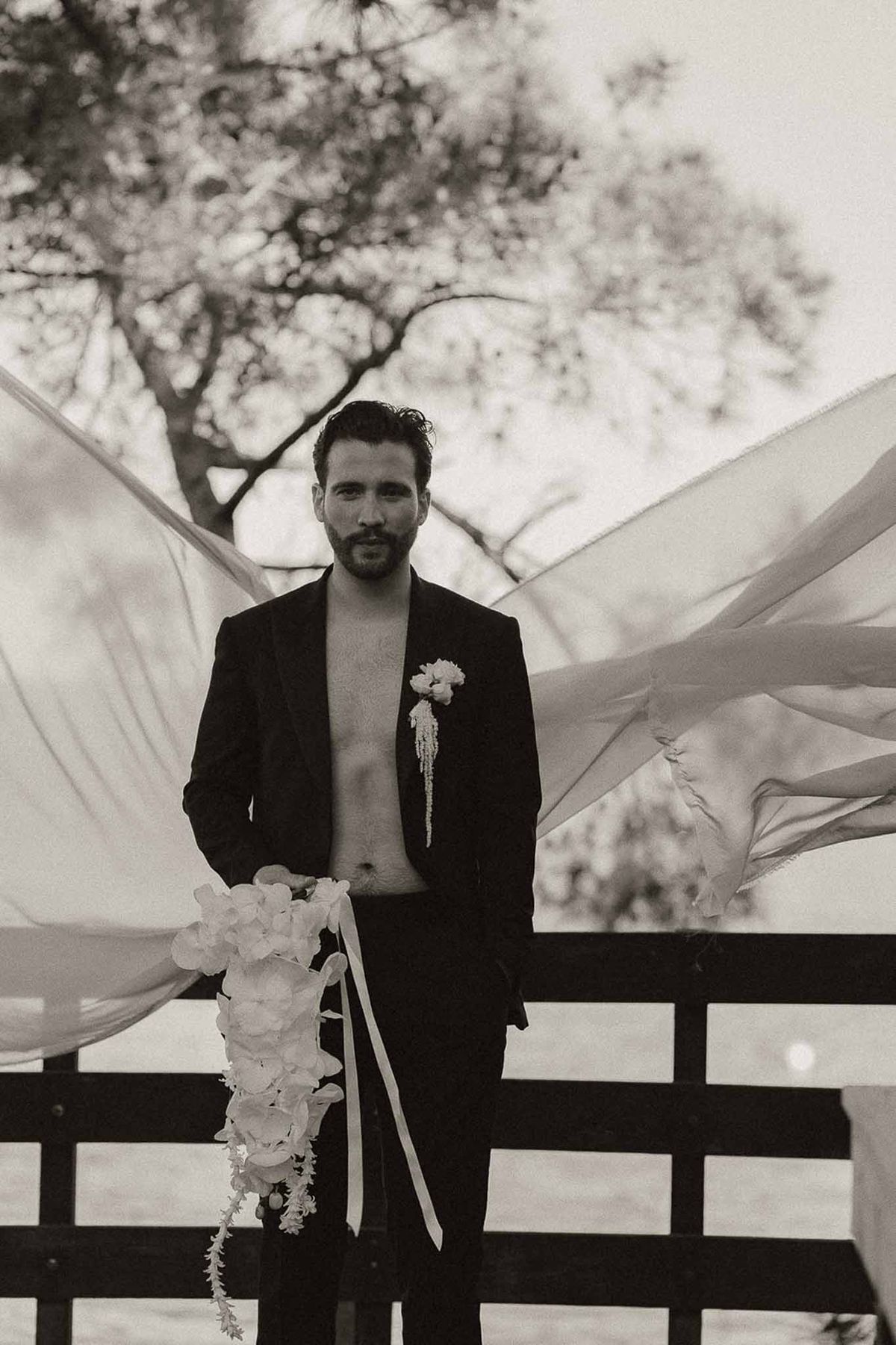 Black and white portrait of groom in open jacket holding cascading hydrangea bouquet with ribbon streamers