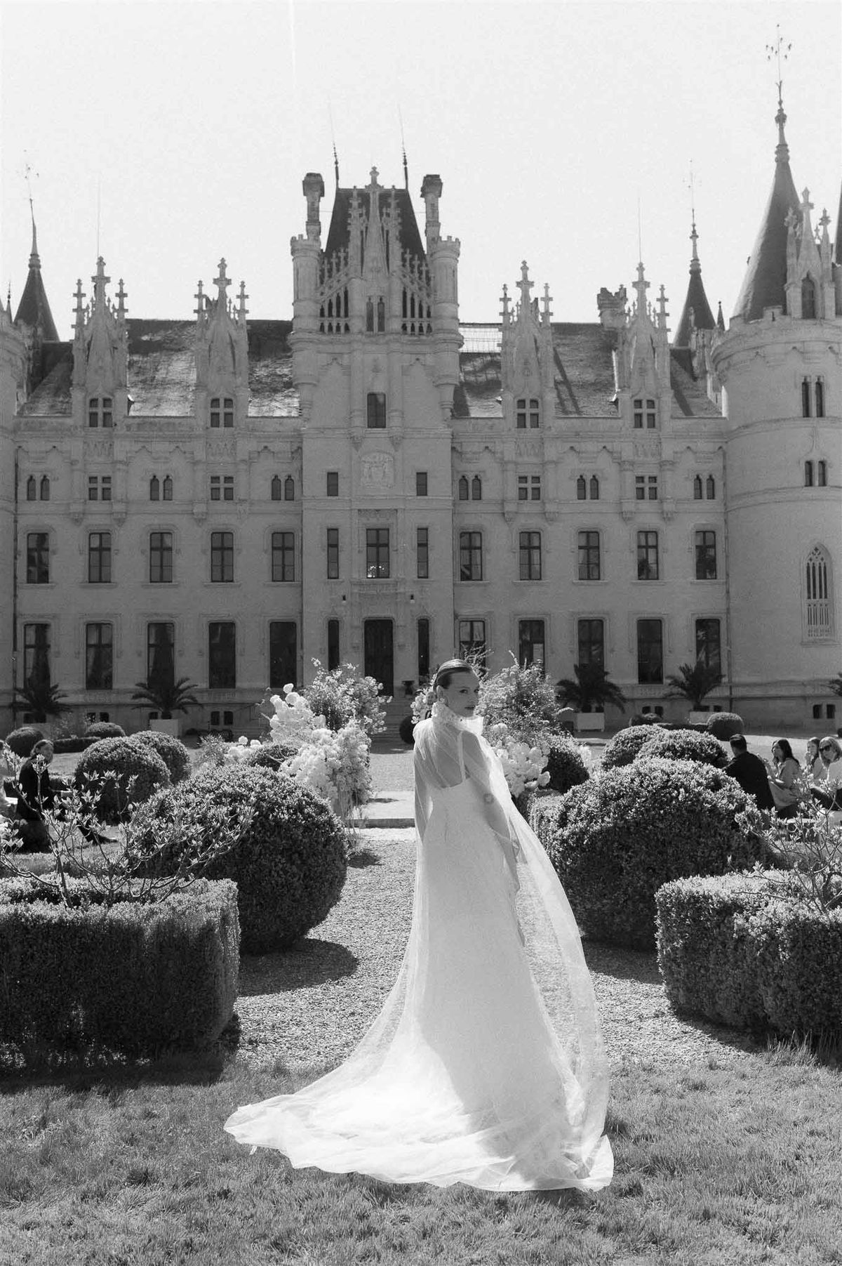 Bride in long-sleeve gown with sweeping veil on parterre garden path before Neo-Gothic chateau in B&W