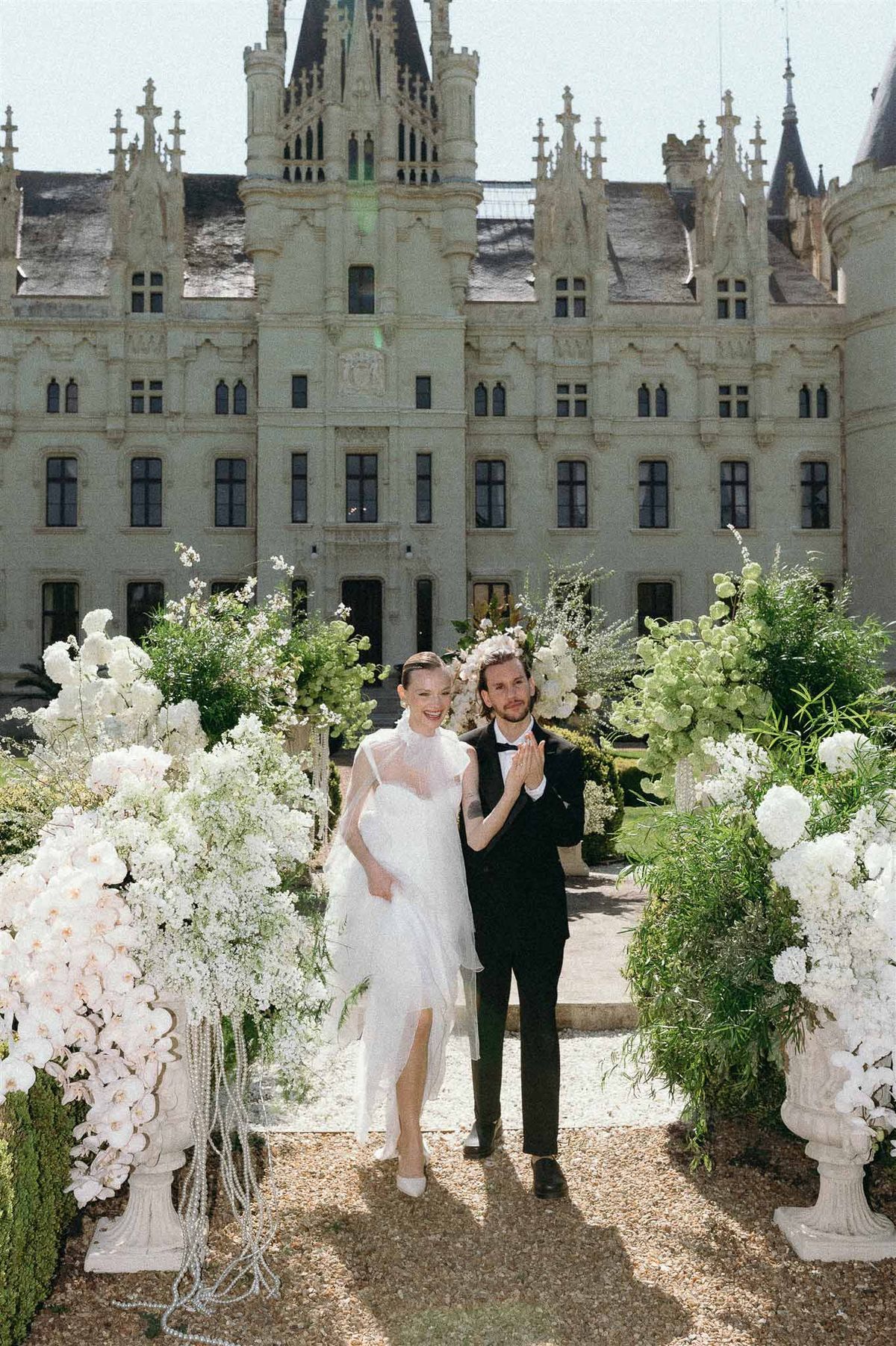 Bride and groom holding hands in chateau courtyard flanked by white orchid and hydrangea arrangements