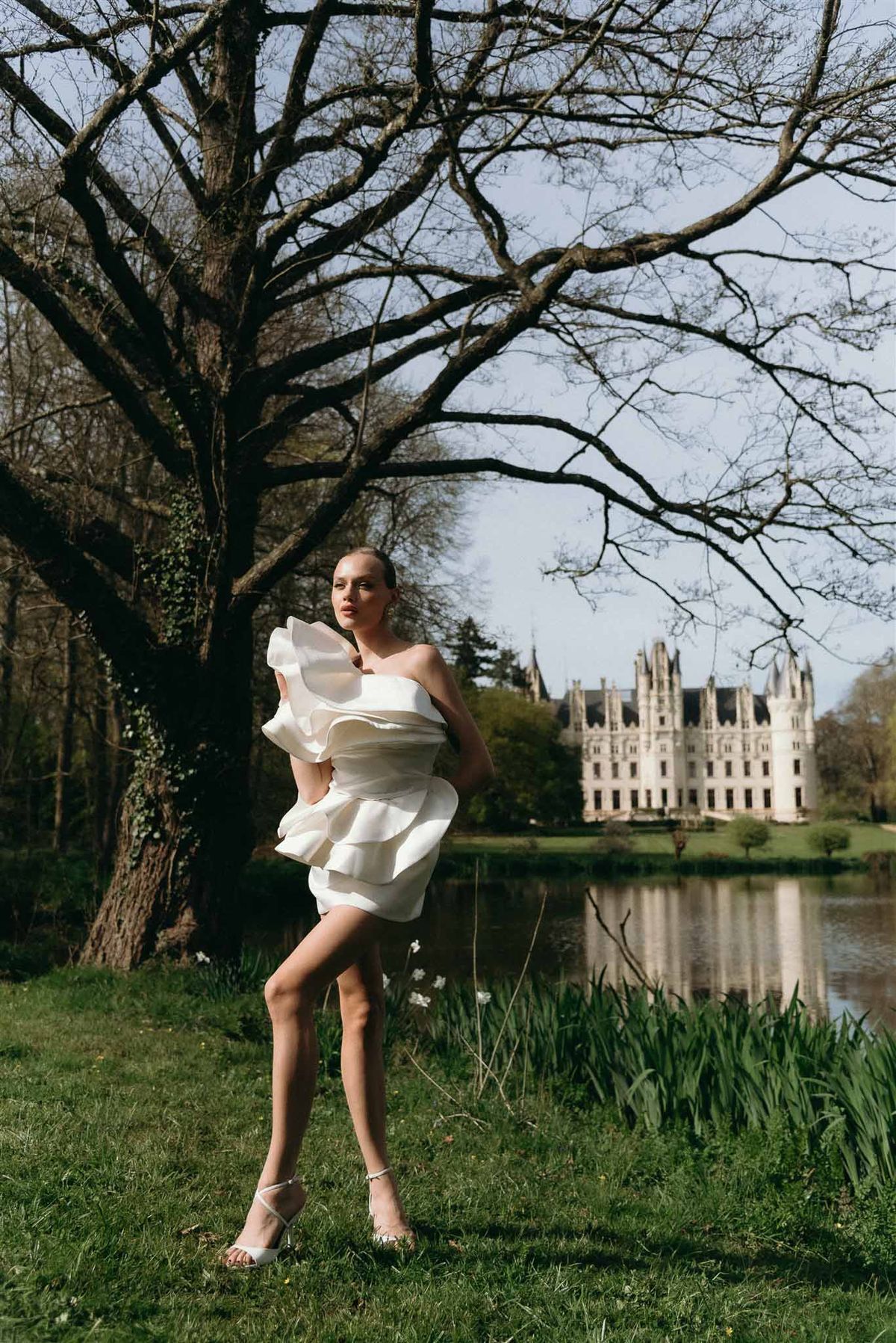 Bride in short ivory ruffled mini dress posing on chateau lawn beside lake with castle reflection