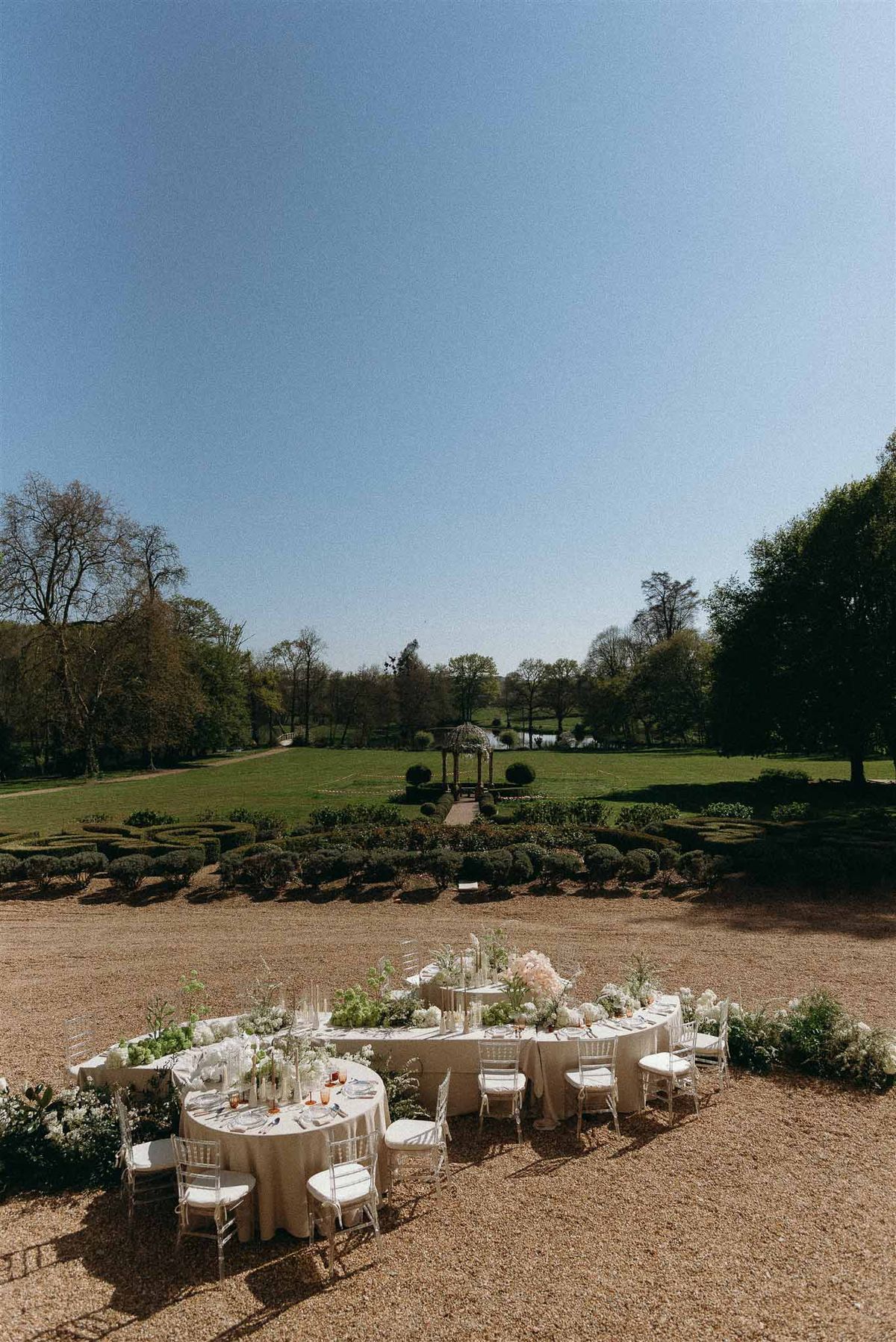 Round and long tables with white blooms, pampas grass, amber glassware, and acrylic chairs on gravel terrace