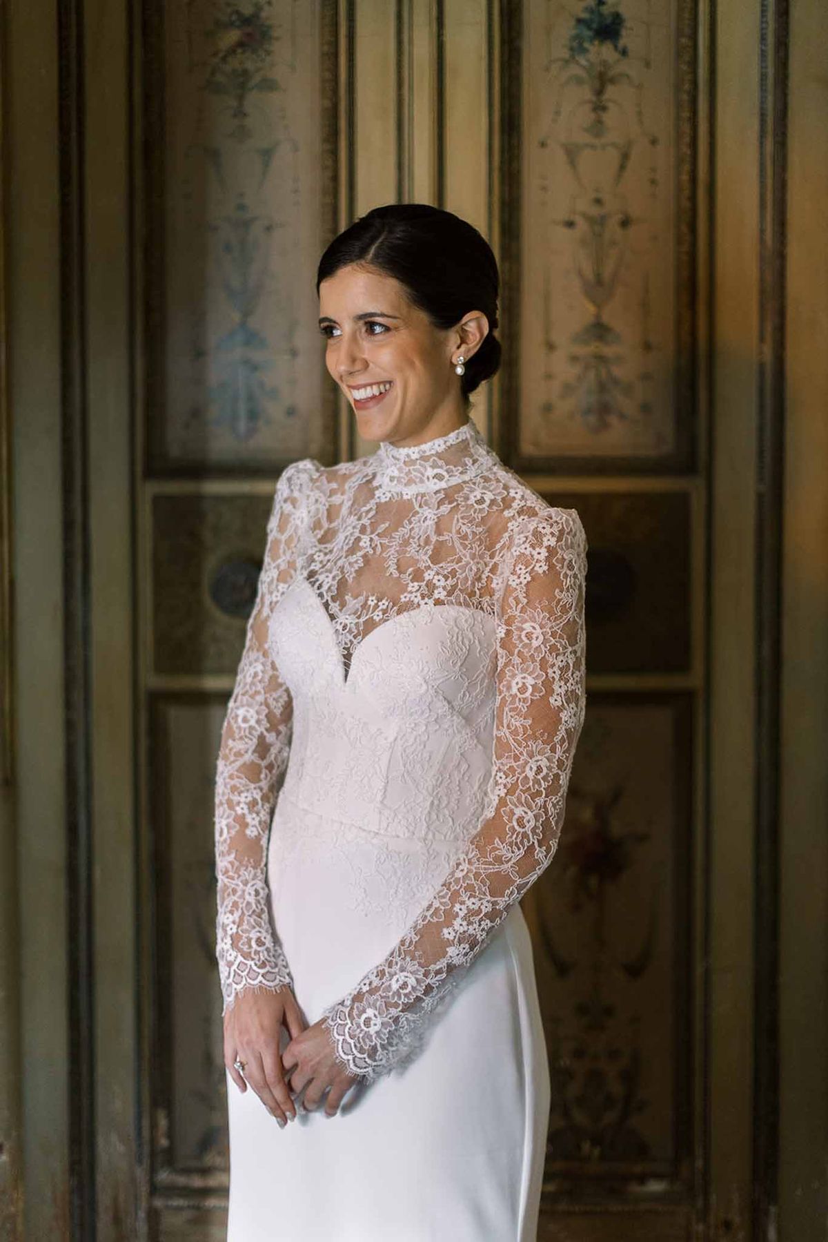 Dark-haired bride in lace high-neck long-sleeve gown smiling in front of painted chateau wall panels