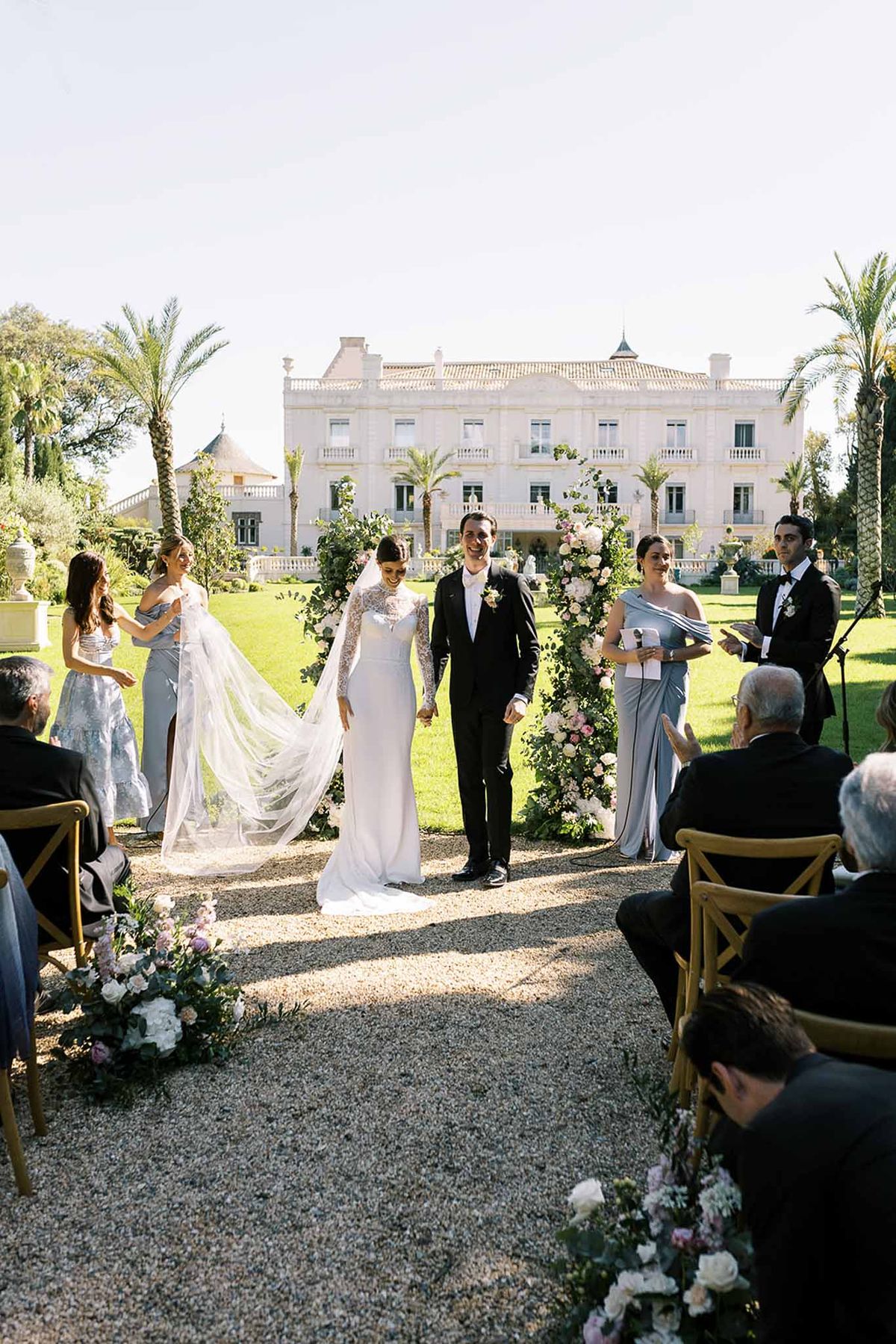 Couple exits greenery and rose arch as bridesmaid holds veil with dusty blue party before Belle Epoque manor