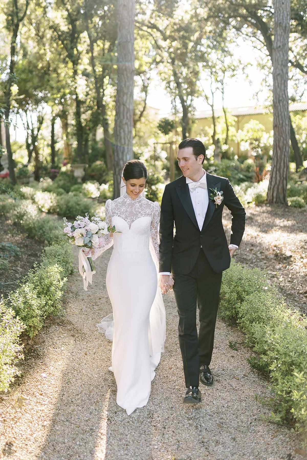 Couple walking on garden path, bride in ivory lace gown with blush peony bouquet, groom in black tuxedo with white bow tie