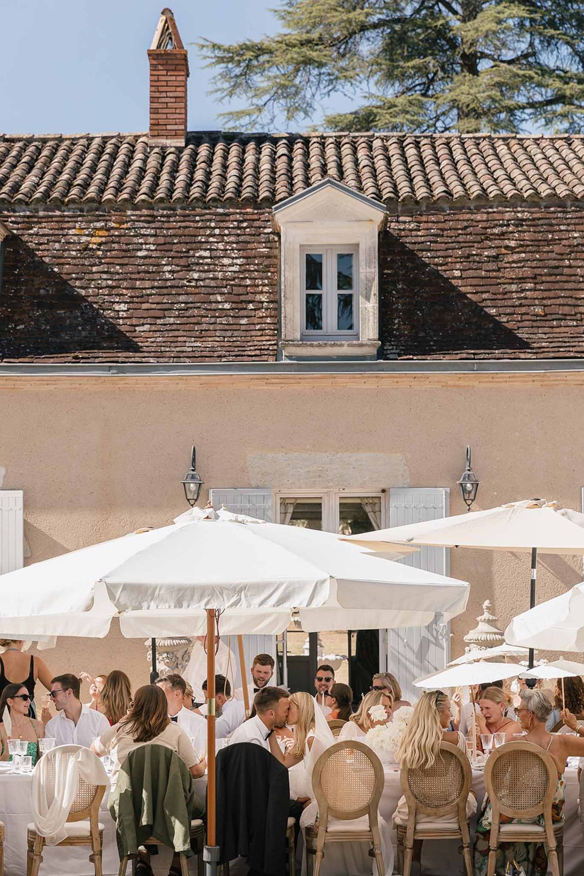 Couple kissing among 25 guests at long table with white umbrellas before terracotta-roofed manor