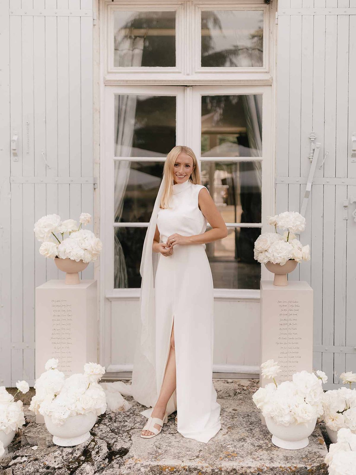 Bride in ivory high-neck cape gown standing among white hydrangea and rose urns outside grey French doors