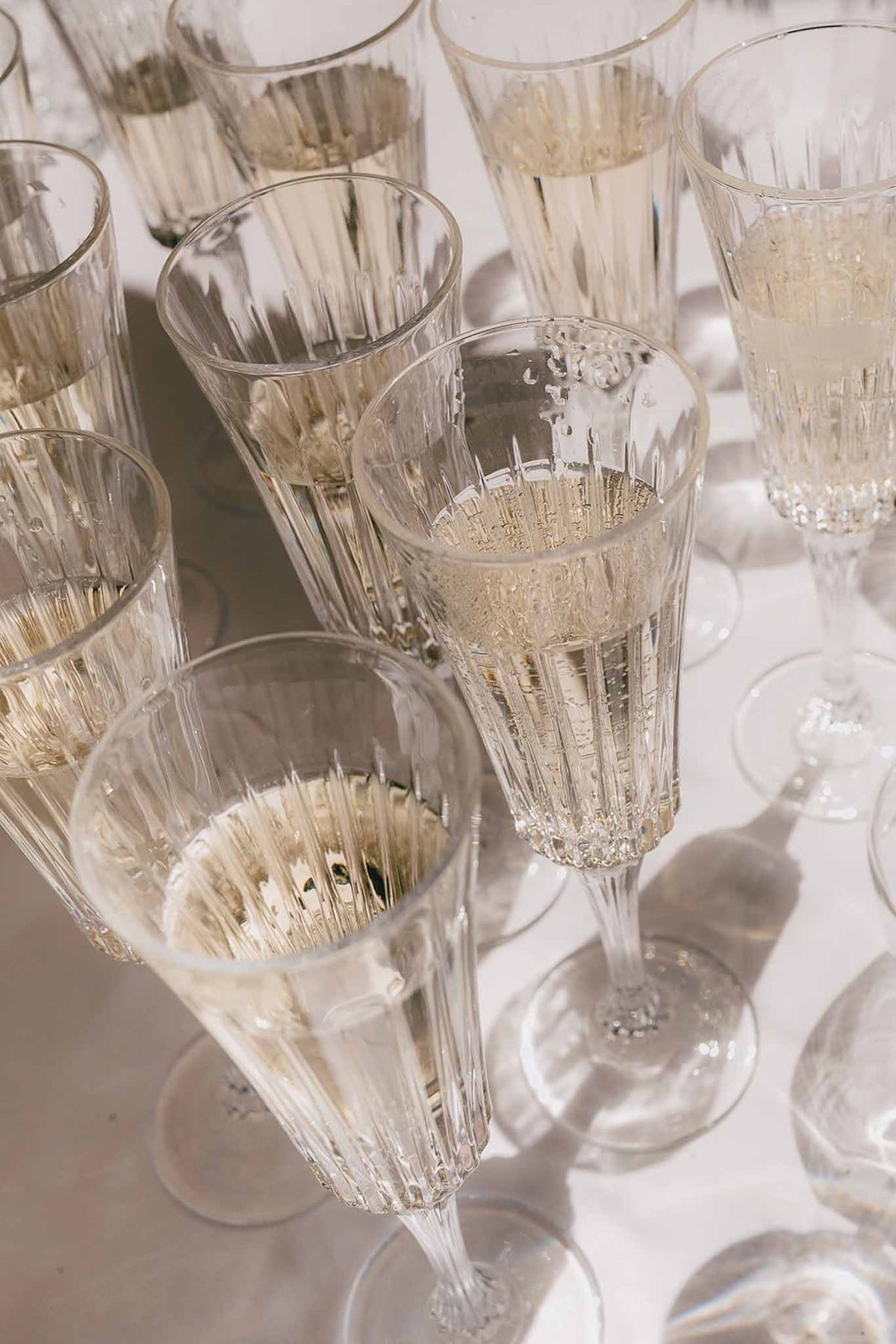 Ten crystal champagne flutes filled with sparkling wine arranged on white linen surface