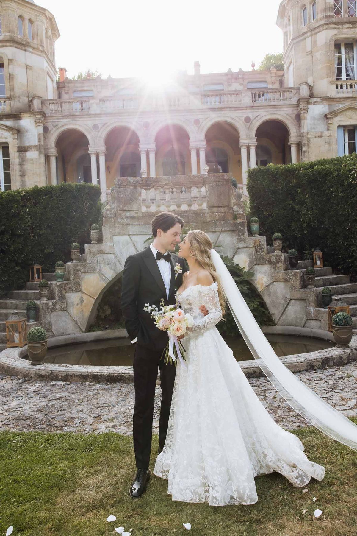 Couple in sunlit colonnade courtyard with stone fountain, cathedral veil trailing behind bride
