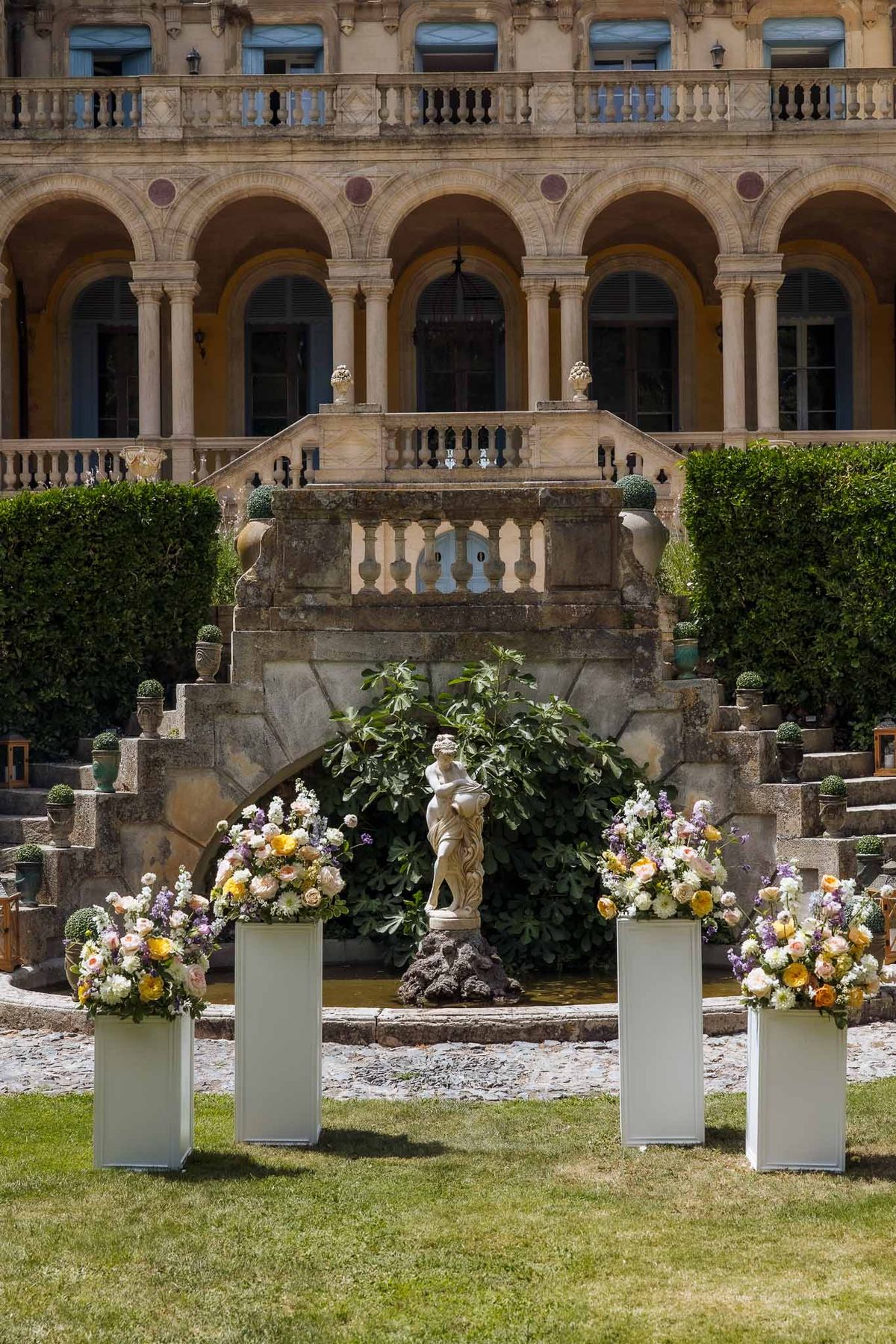 Ceremony pedestals with peach and lavender florals before classical chateau colonnade and fountain