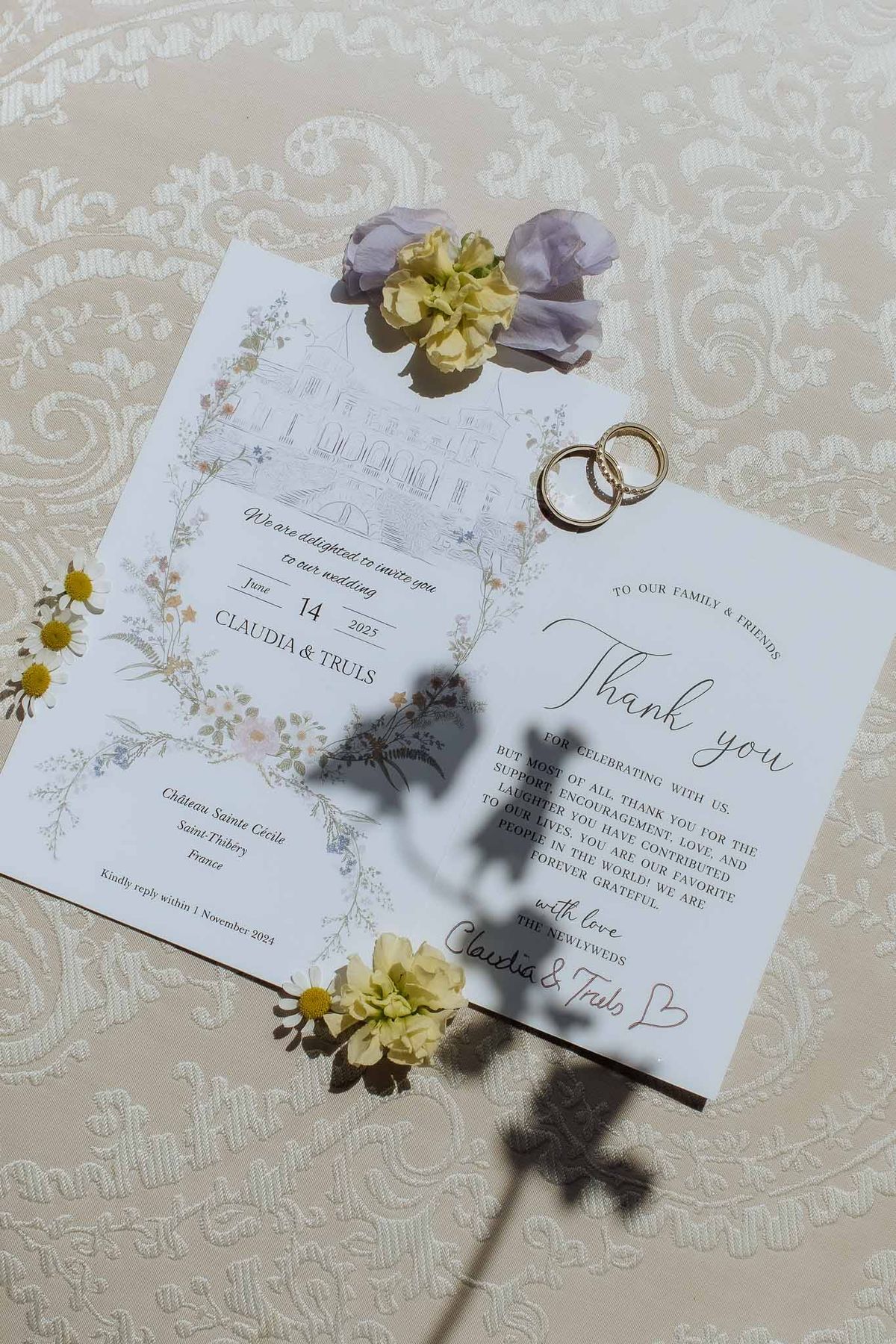 Chateau illustration invitation with wildflower border, gold wedding bands, and loose daisies on jacquard