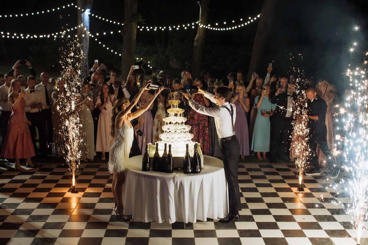 Couple pouring champagne tower on checkered dance floor with sparkler fountains and guests filming at evening reception