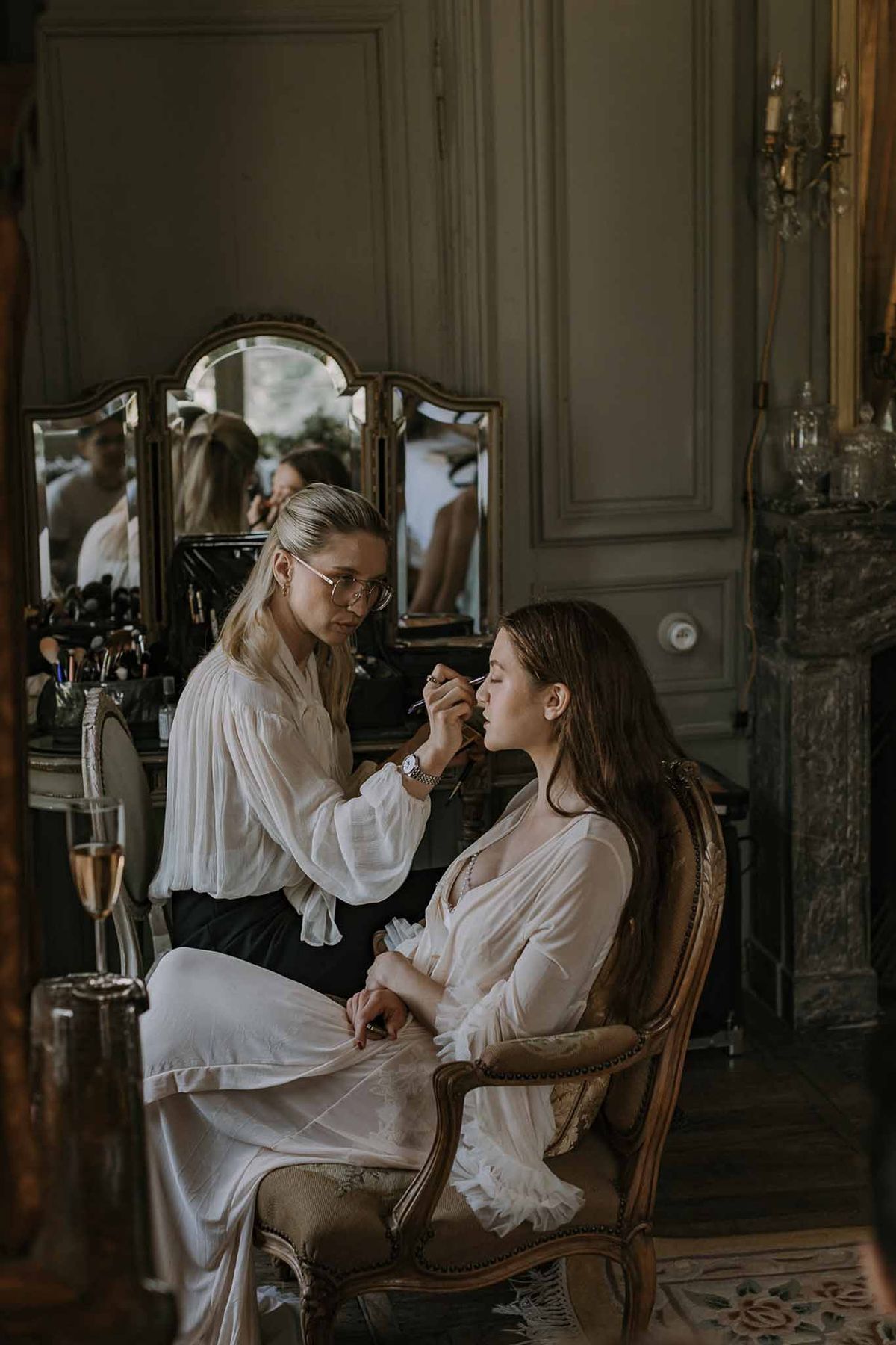 Makeup artist applies lipstick to bride in gilt armchair before tri-fold vanity in grey-panelled chateau room