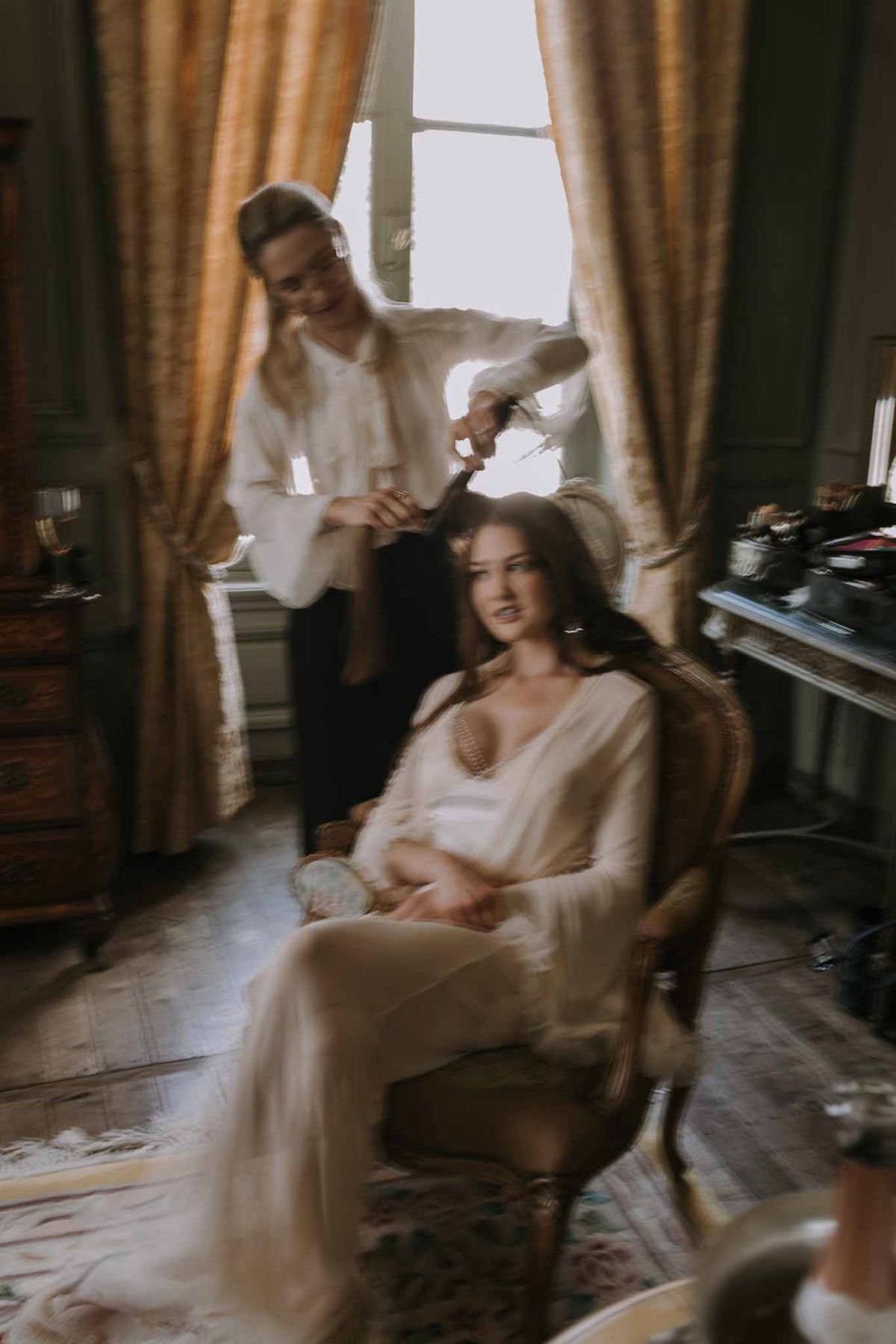 Motion-blurred getting ready scene of hairstylist working on bride in ivory robe in gold-curtained chateau room