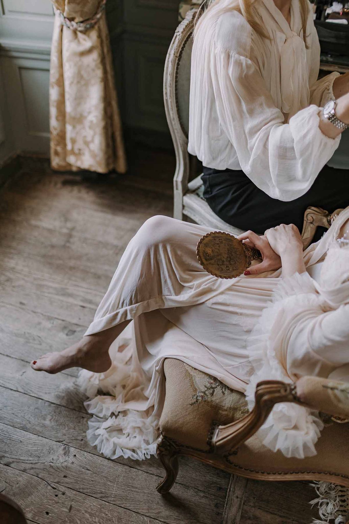 Bride in ivory silk robe with tulle trim holds antique bronze mirror on gilded armchair in chateau room