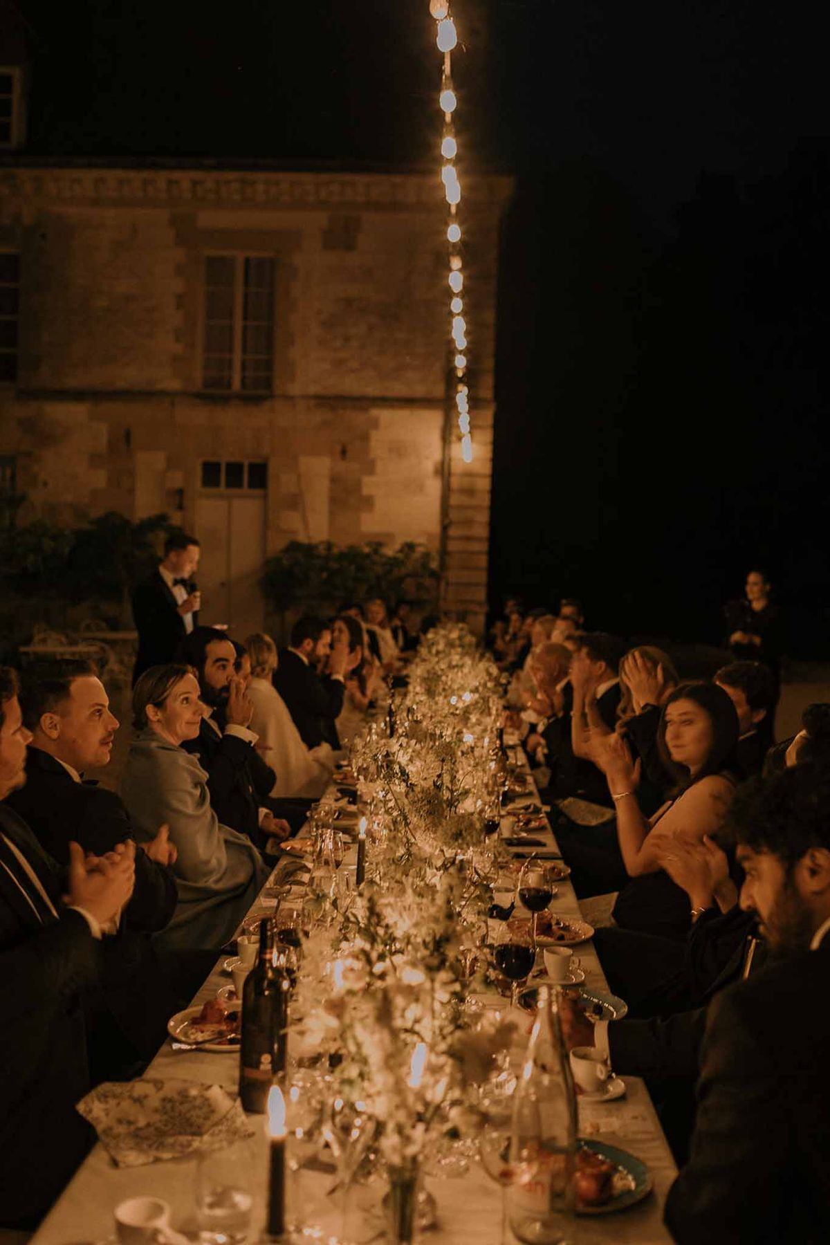 Evening reception speech at long candlelit table with vertical fairy lights in chateau courtyard