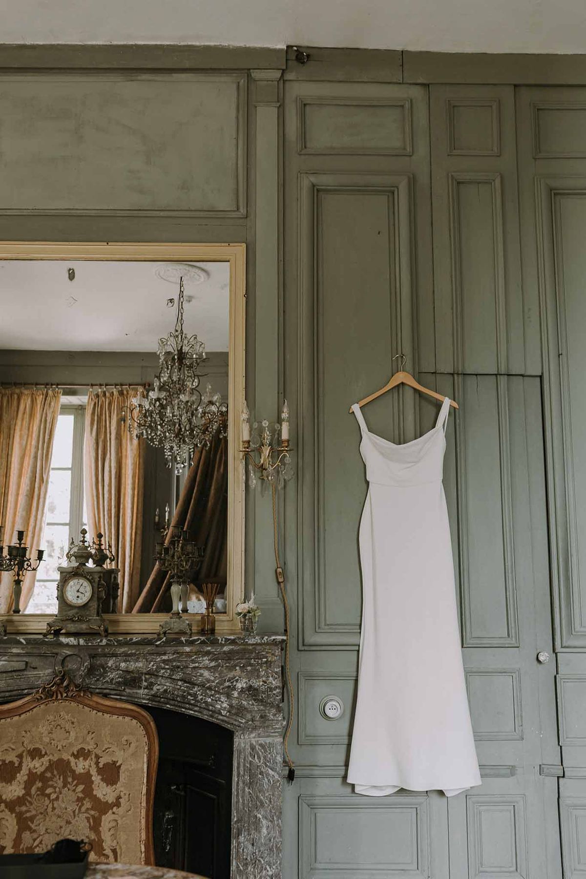 Ivory slip wedding dress on hanger beside grey marble fireplace with gilt mirror in sage-green chateau bedroom