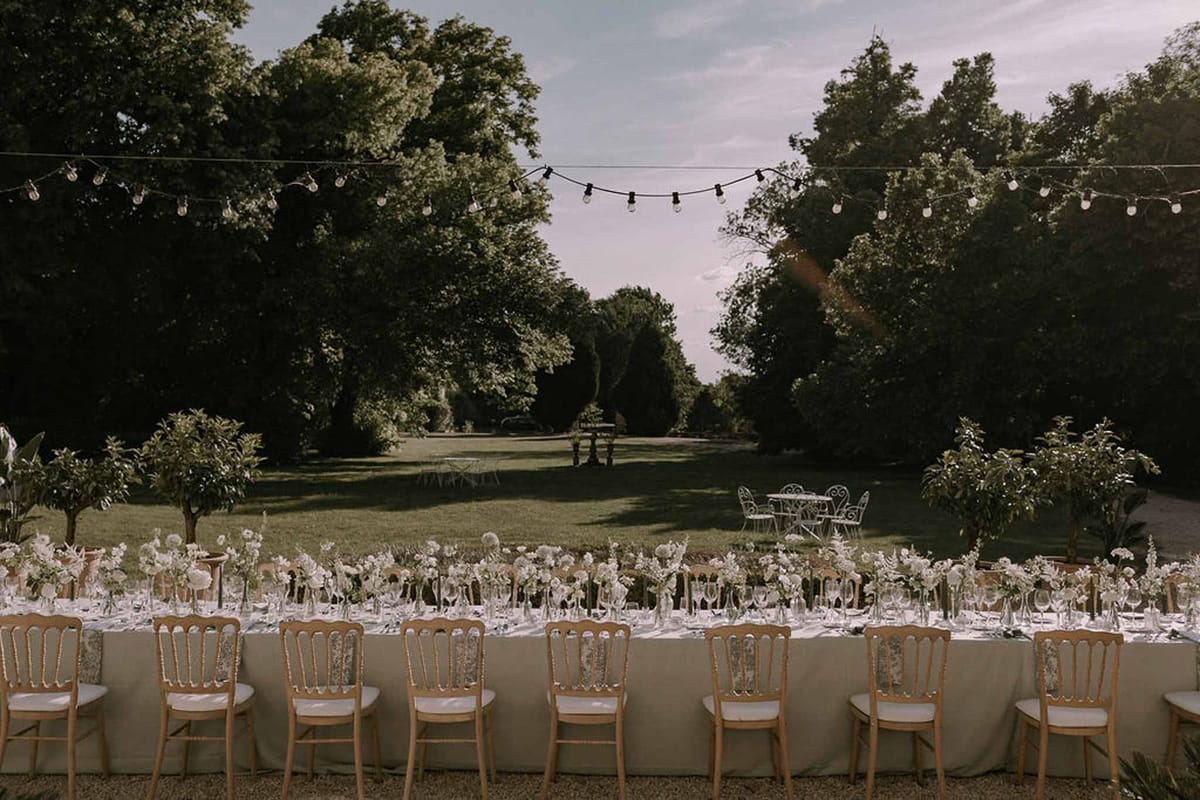 Long banquet table with peony and allium bud vases, Napoleon chairs, and Edison lights in garden
