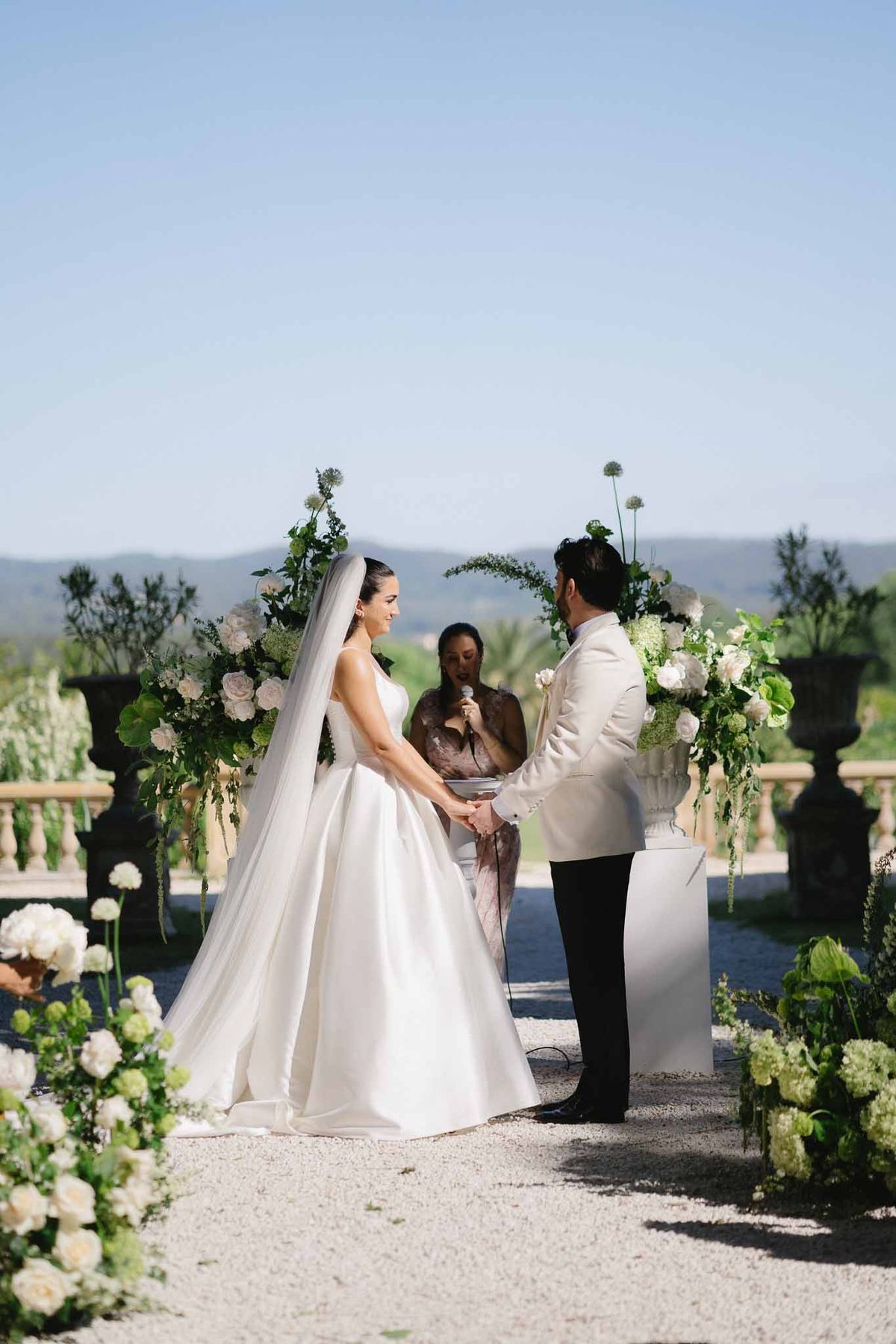Bride and groom holding hands at an outdoor ceremony on a hillside terrace with a circular floral arch and stone urns