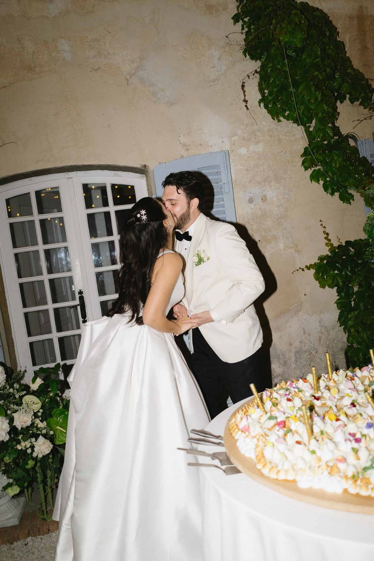 Couple kisses at cake cutting beside white anthurium arrangement against vine-covered chateau wall