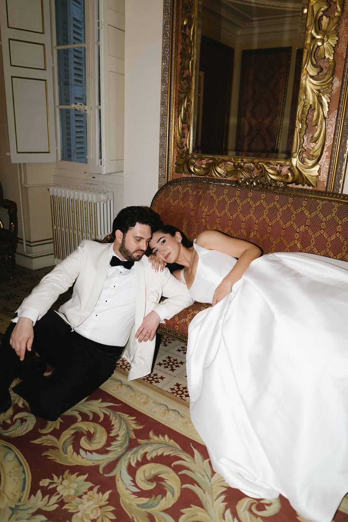 Groom on floor and bride reclining on antique red sofa before gilt baroque mirror in chateau salon