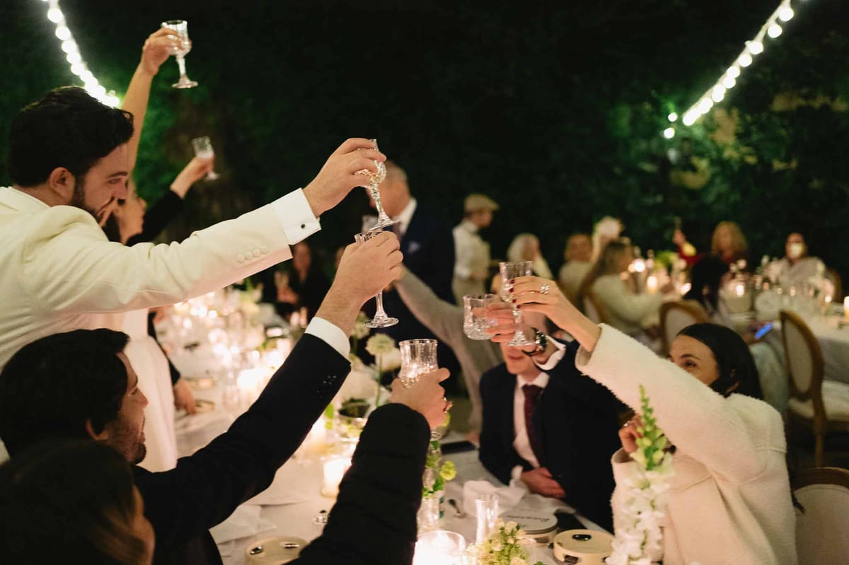 Guests raising crystal glasses in toast at outdoor evening reception with string lights and candlelit tables