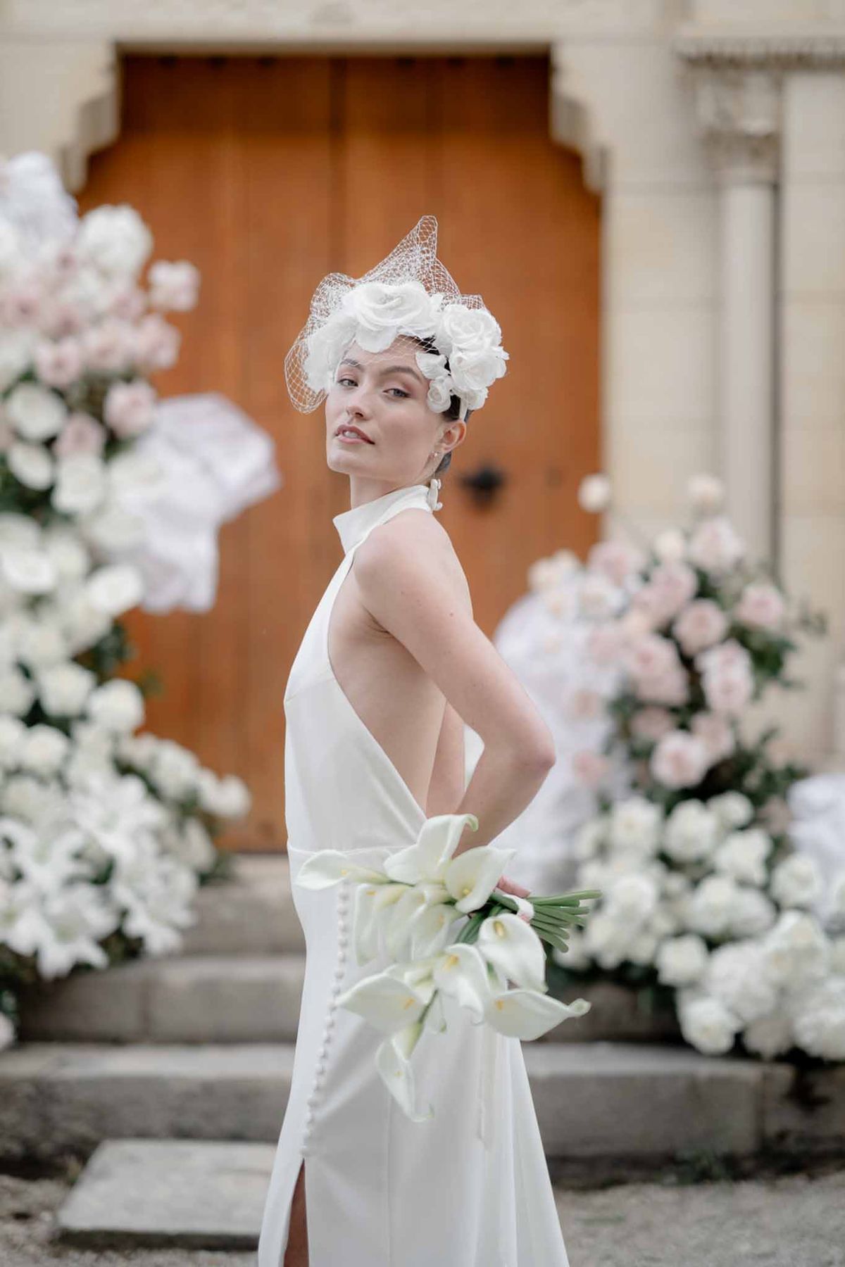 Bride in white halter-neck open-back gown with birdcage fascinator and calla lily bouquet beside rose installations