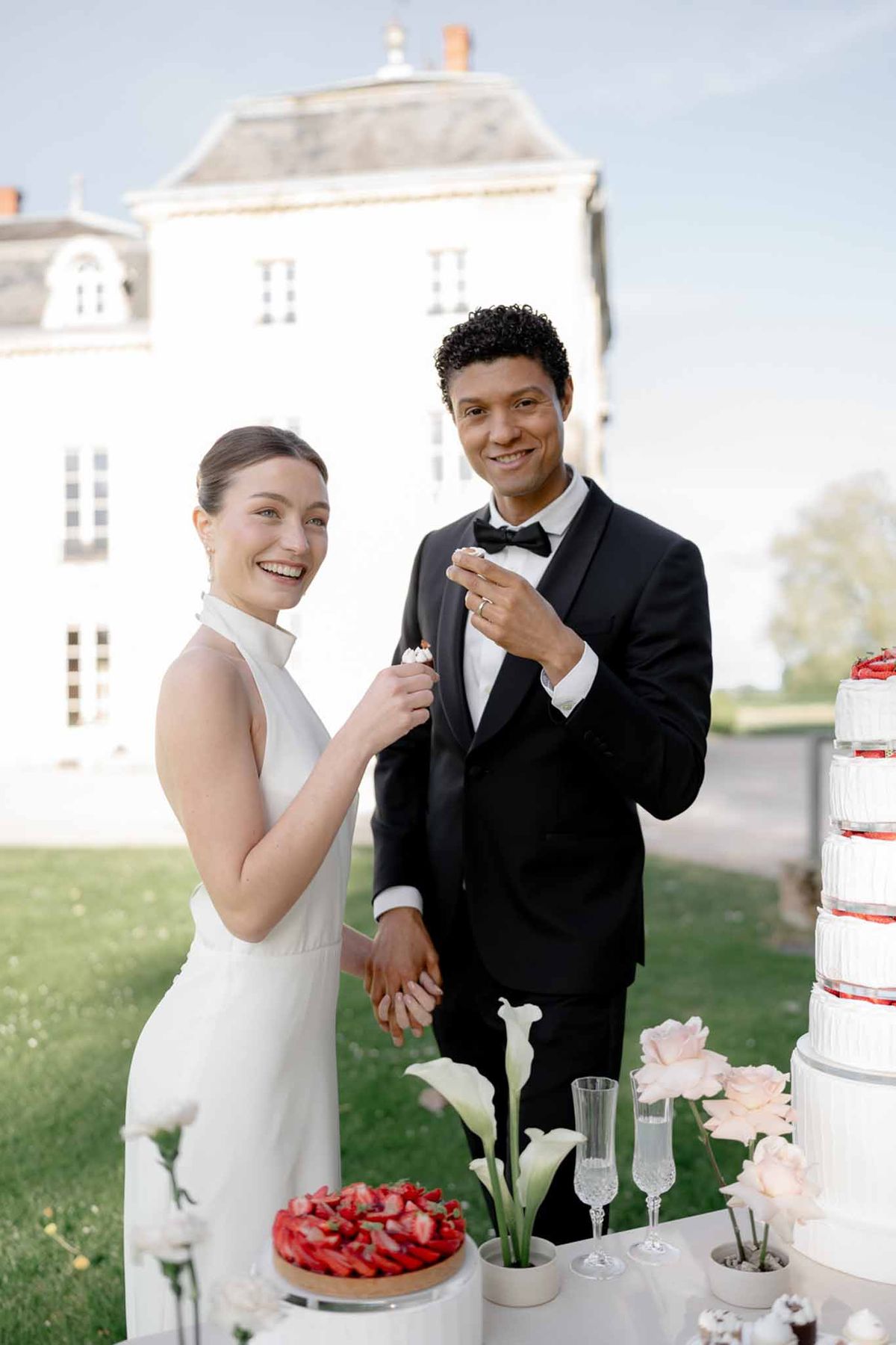 Couple cuts ribbed white cake with fresh strawberries and calla lilies before white chateau facade