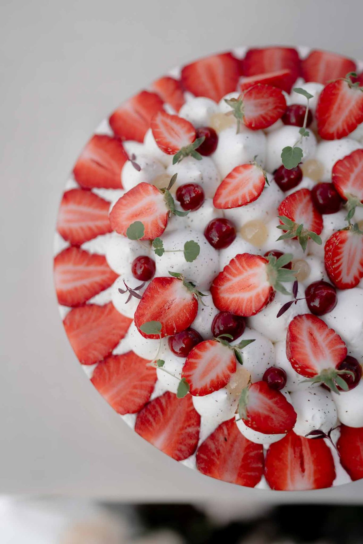 Top of wedding cake with white meringue texture, halved strawberries, dark cherries, and fresh herb sprigs