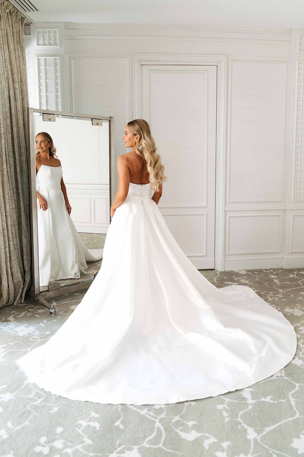 Bride seen from behind in strapless white satin ball gown with cathedral train, looking into freestanding mirror