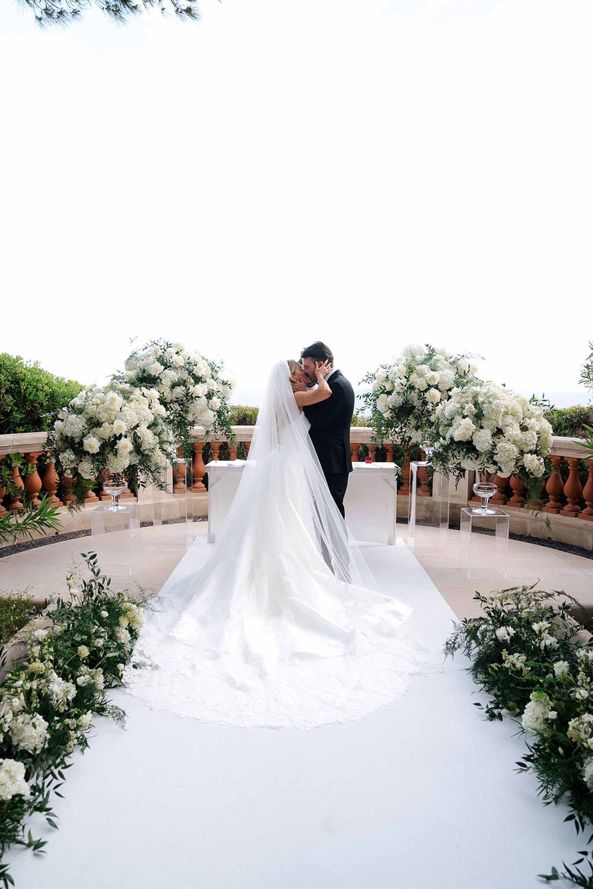 First kiss on balustrade terrace with white rose pedestals and cathedral veil spread on aisle runner