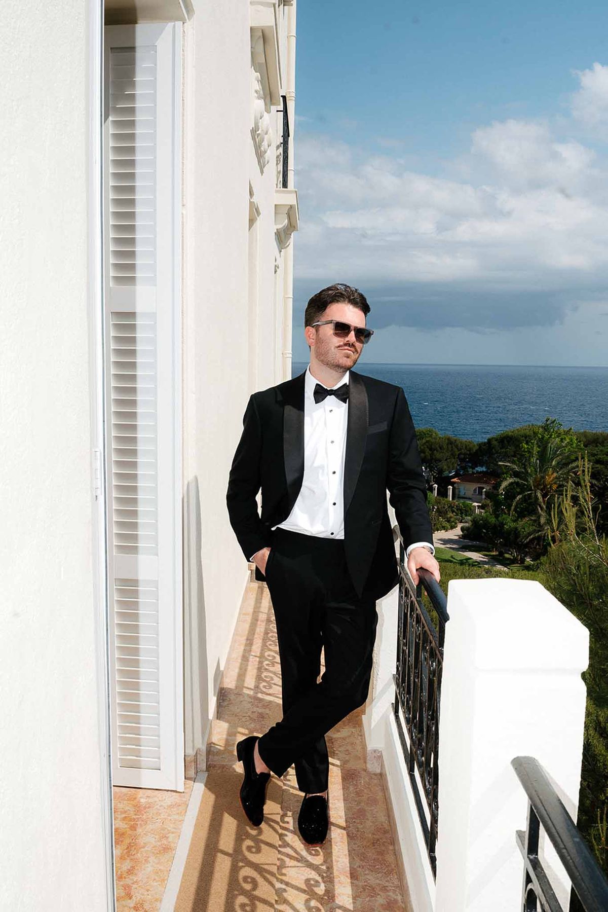 Groom in black tuxedo and sunglasses leaning on a balcony railing with sea view in background
