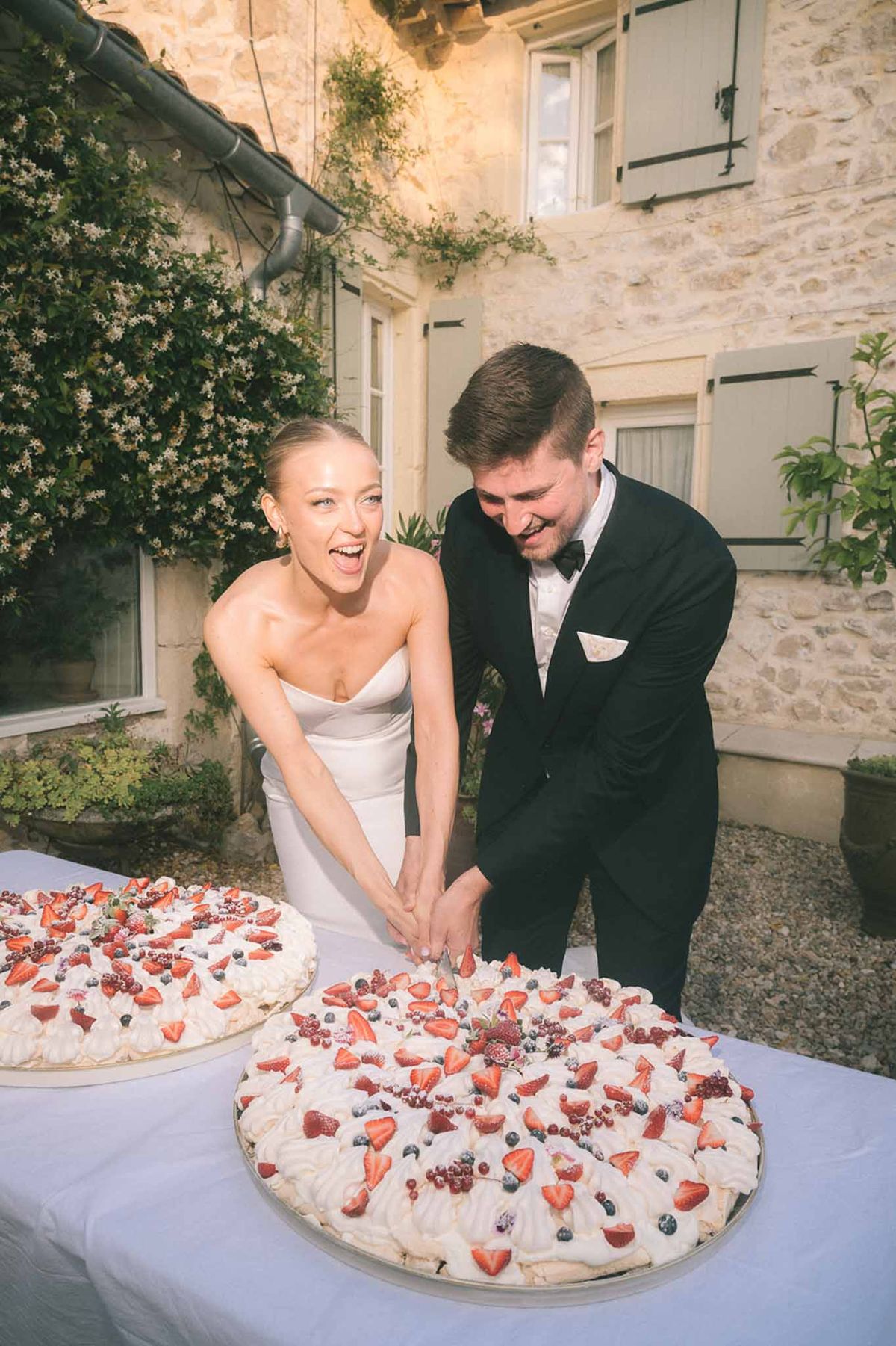 Laughing couple cutting pavlovas topped with fresh berries outdoors at stone-facade venue