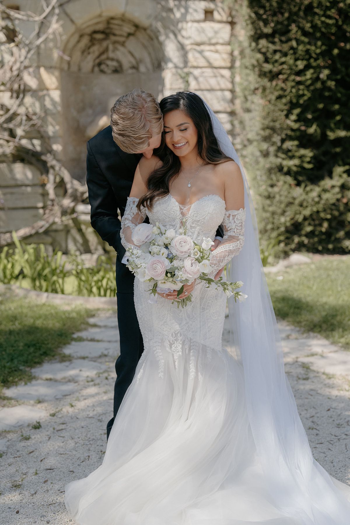 Bride in ivory lace mermaid gown with cathedral veil and blush bouquet as groom leans to her cheek